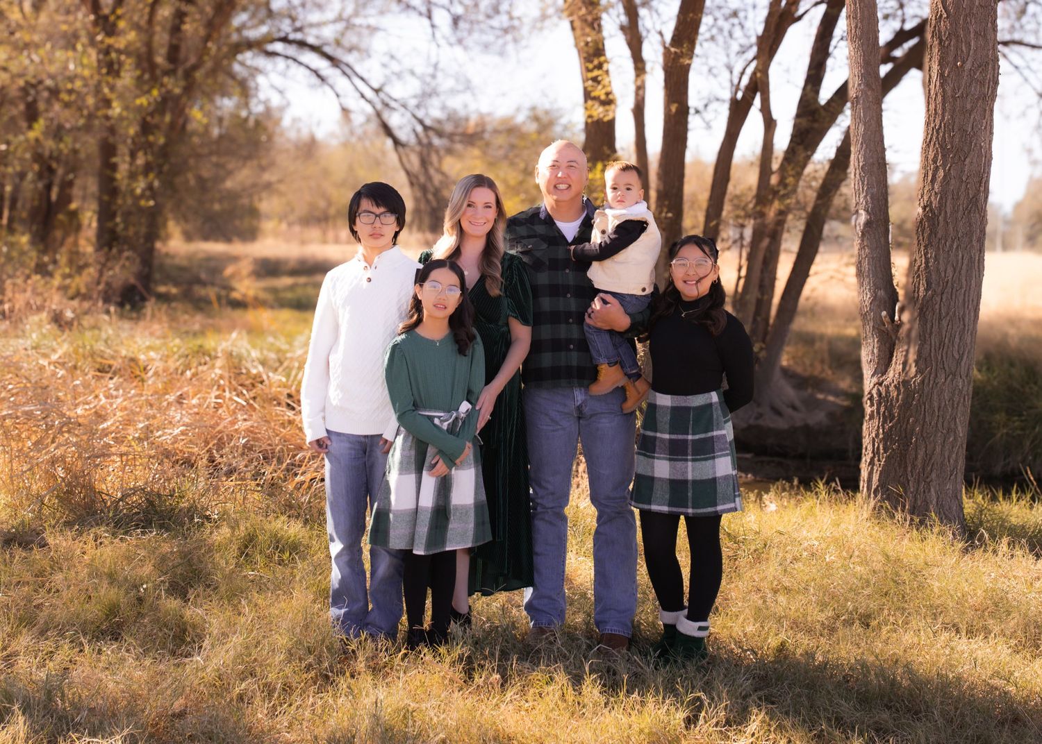 Family of six posing outdoors in autumn woodland setting with golden light filtering through bare trees.