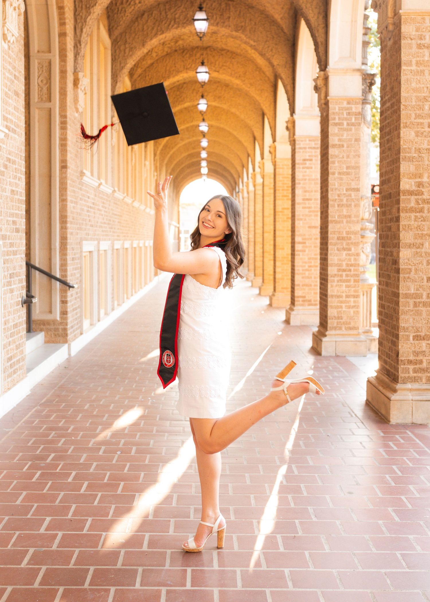 A joyful graduate in a white dress tosses her cap in an arched brick hallway, holding a diploma.