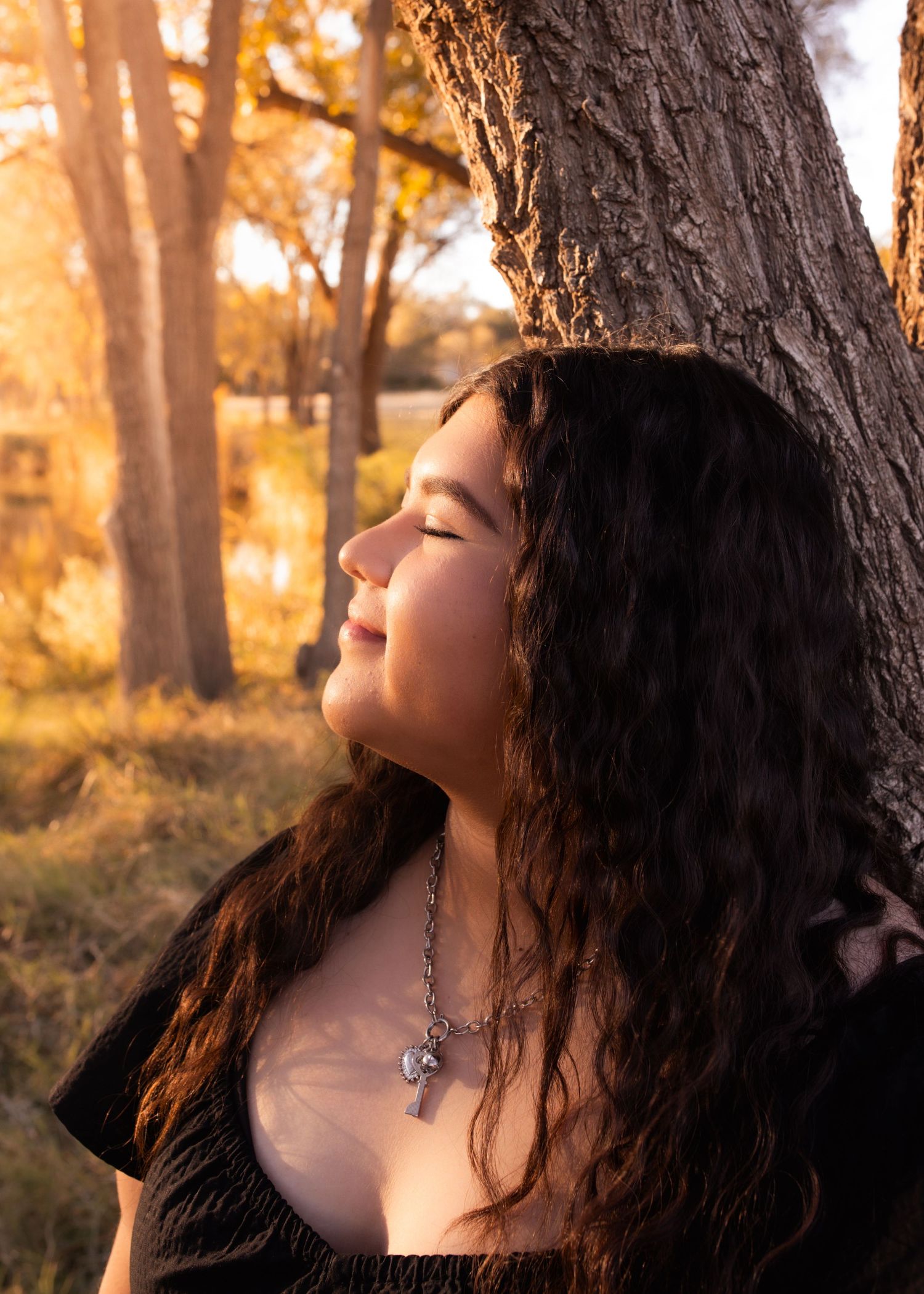 Young woman with curly hair leans against a tree, eyes closed, bathed in warm golden sunset light outdoors.