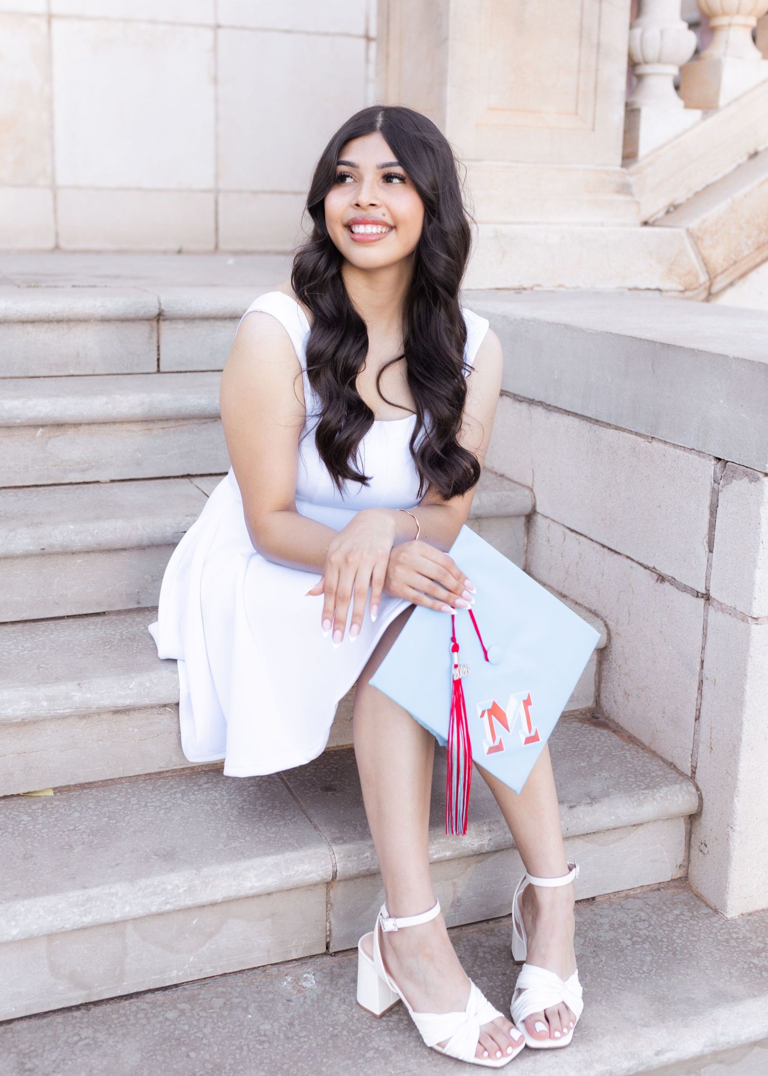 Young woman in white dress sitting on stone steps holding a light blue graduation cap with red tassel, smiling.