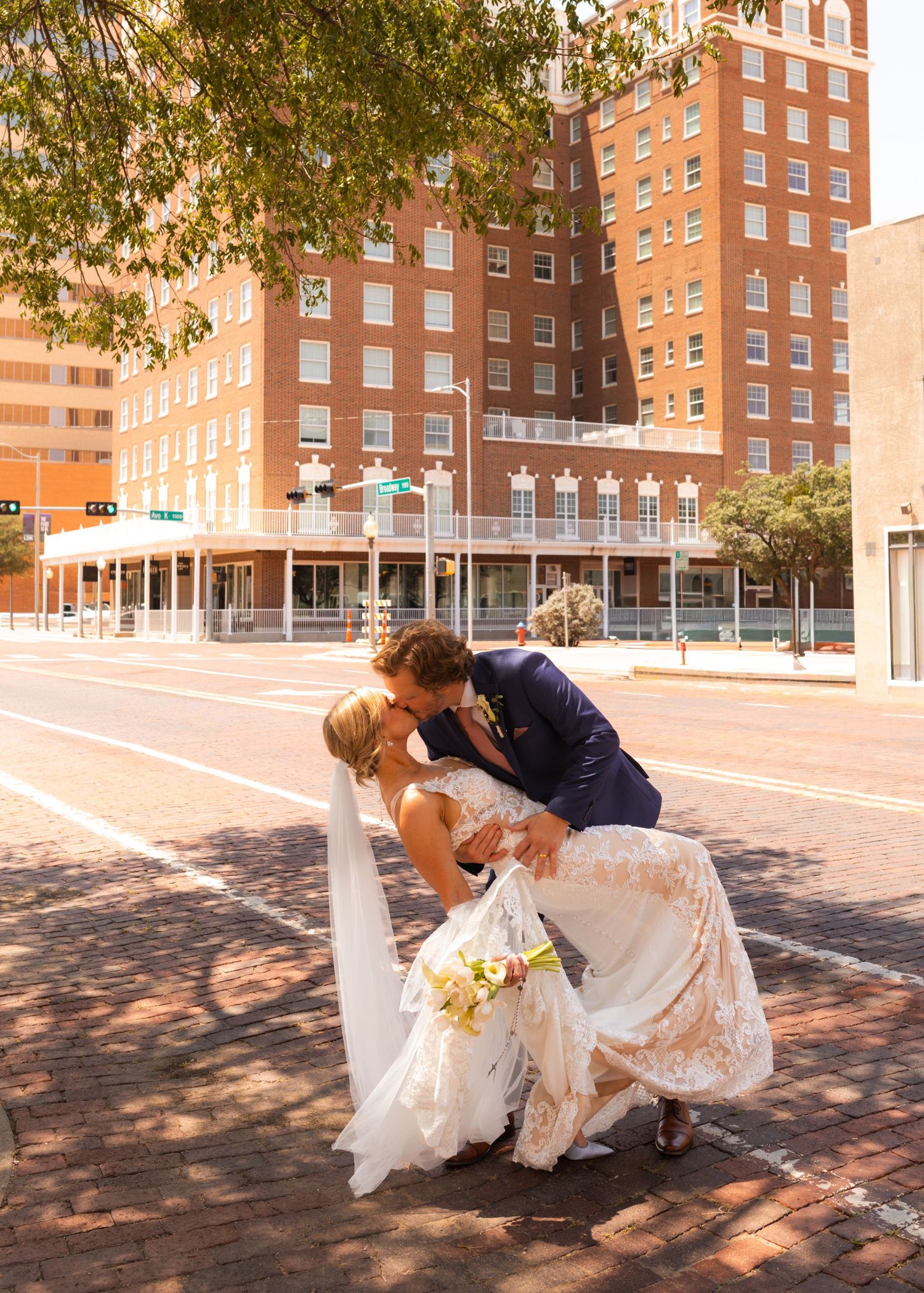 Bride and groom share a romantic dip kiss on a brick sidewalk in front of a historic red brick building.