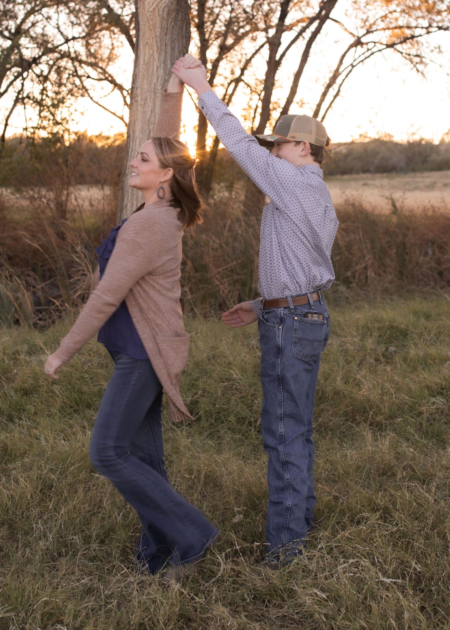 Couple dancing outdoors at sunset near a tree in a rural field, woman in cardigan and man in cowboy hat.