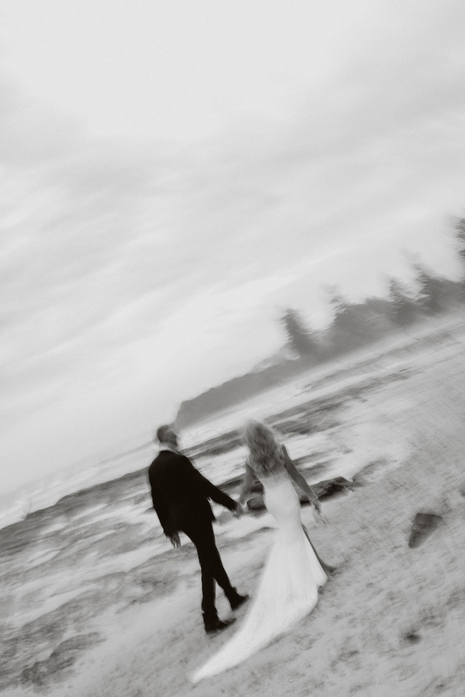 Blurred black and white artistic photo of bride and groom holding hands walking on a windy beach.