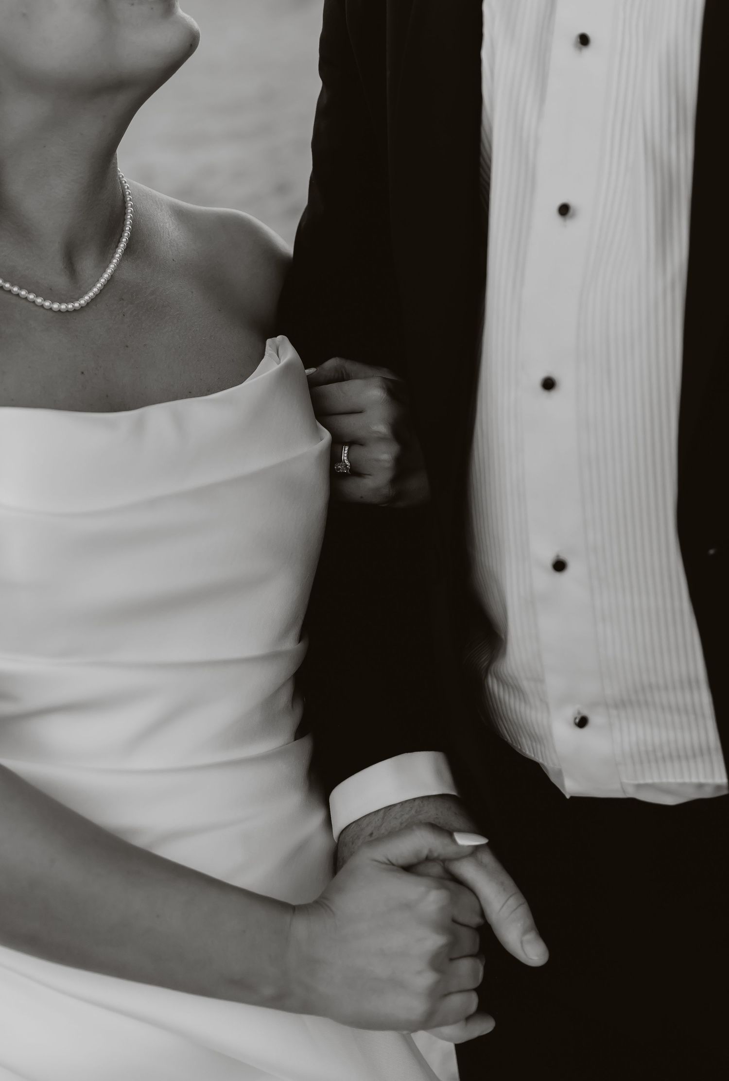 A black and white close-up of a bride in a strapless gown holding her groom's arm and hand tenderly.