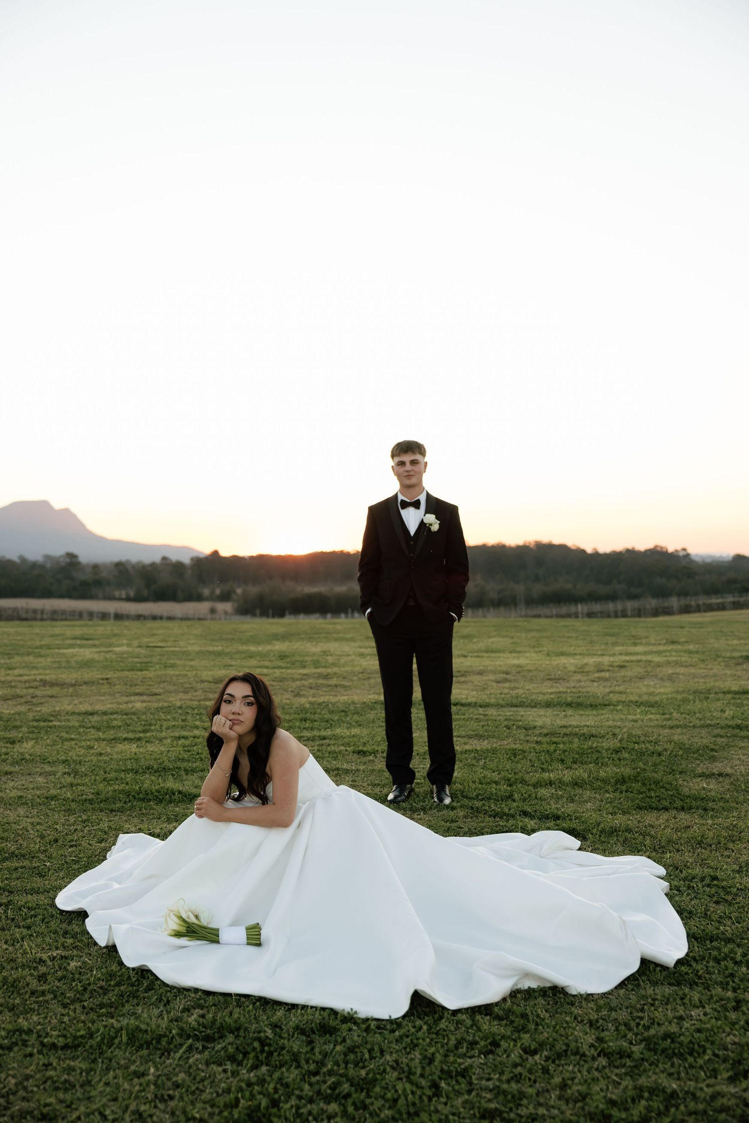 Bride reclines on grass in a flowing white gown as groom stands behind her at a sunset wedding at Bimbadgen Estate.