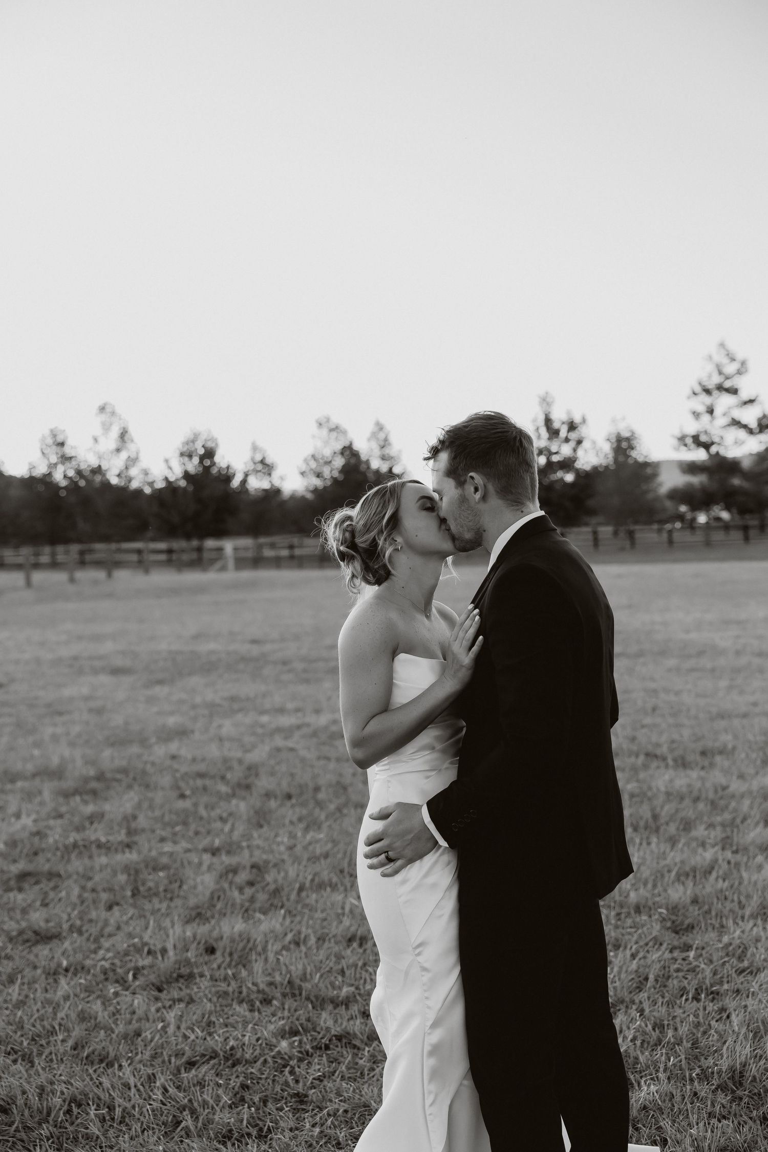 Black and white photo of bride and groom sharing a romantic kiss in an open field at dusk.