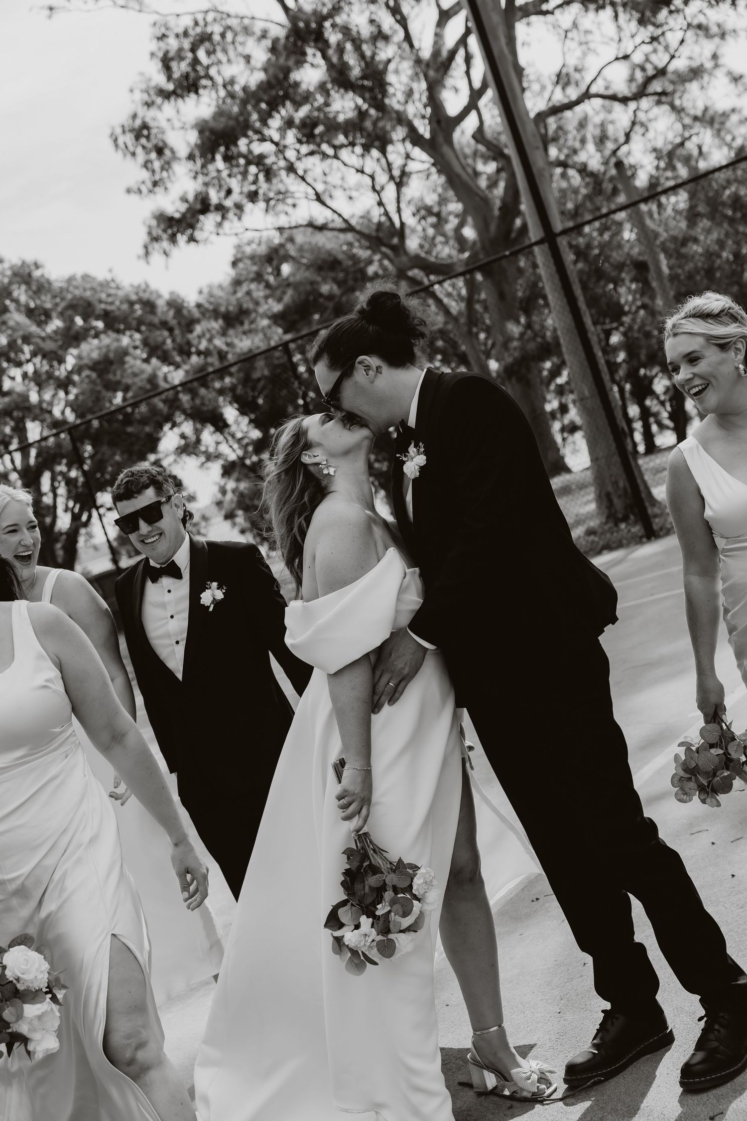 Black and white photo of bride and groom sharing a kiss while wedding party walks alongside them.