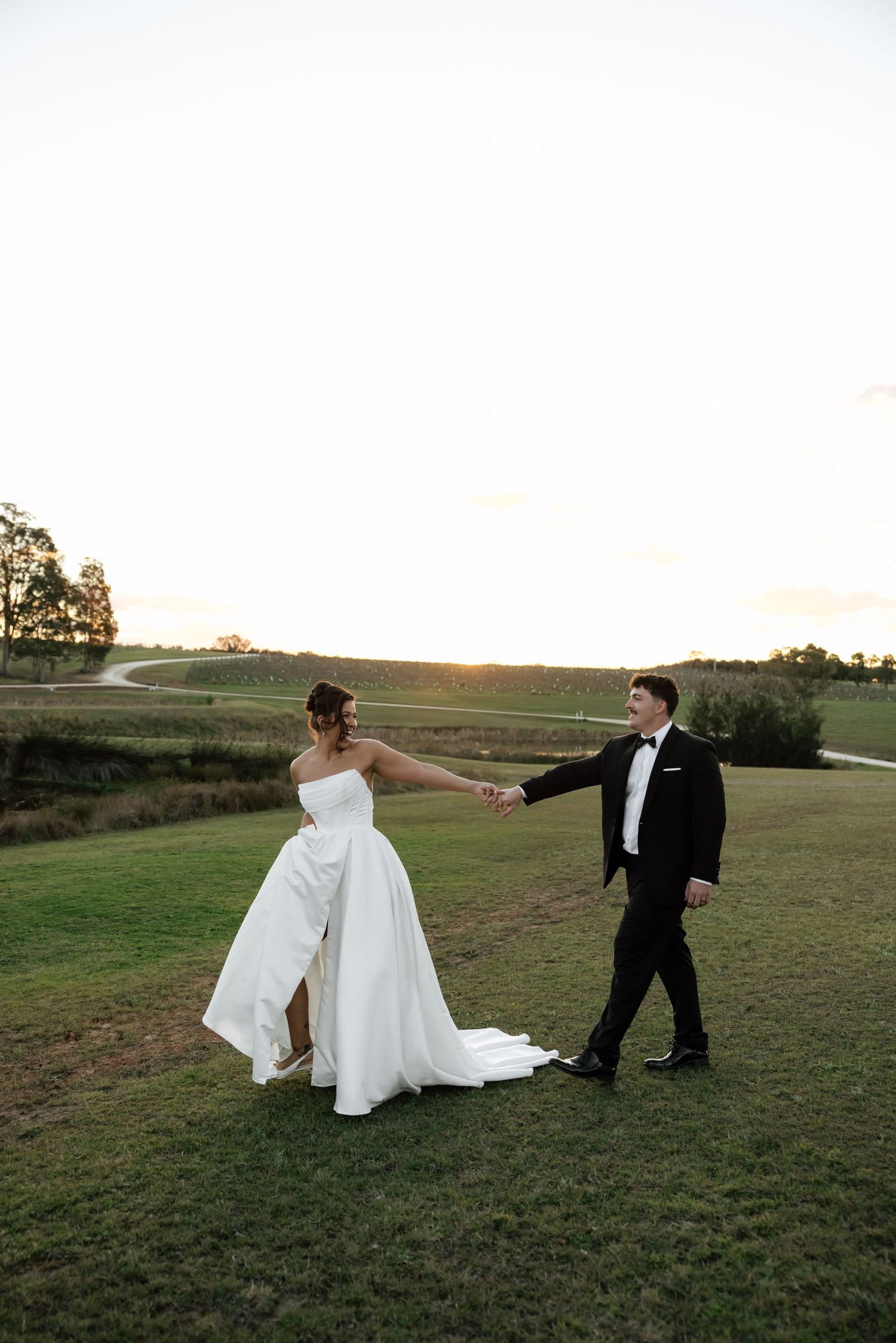 Bride and groom hold hands walking through a scenic green field at golden hour on their wedding day and Squires Vineyard.