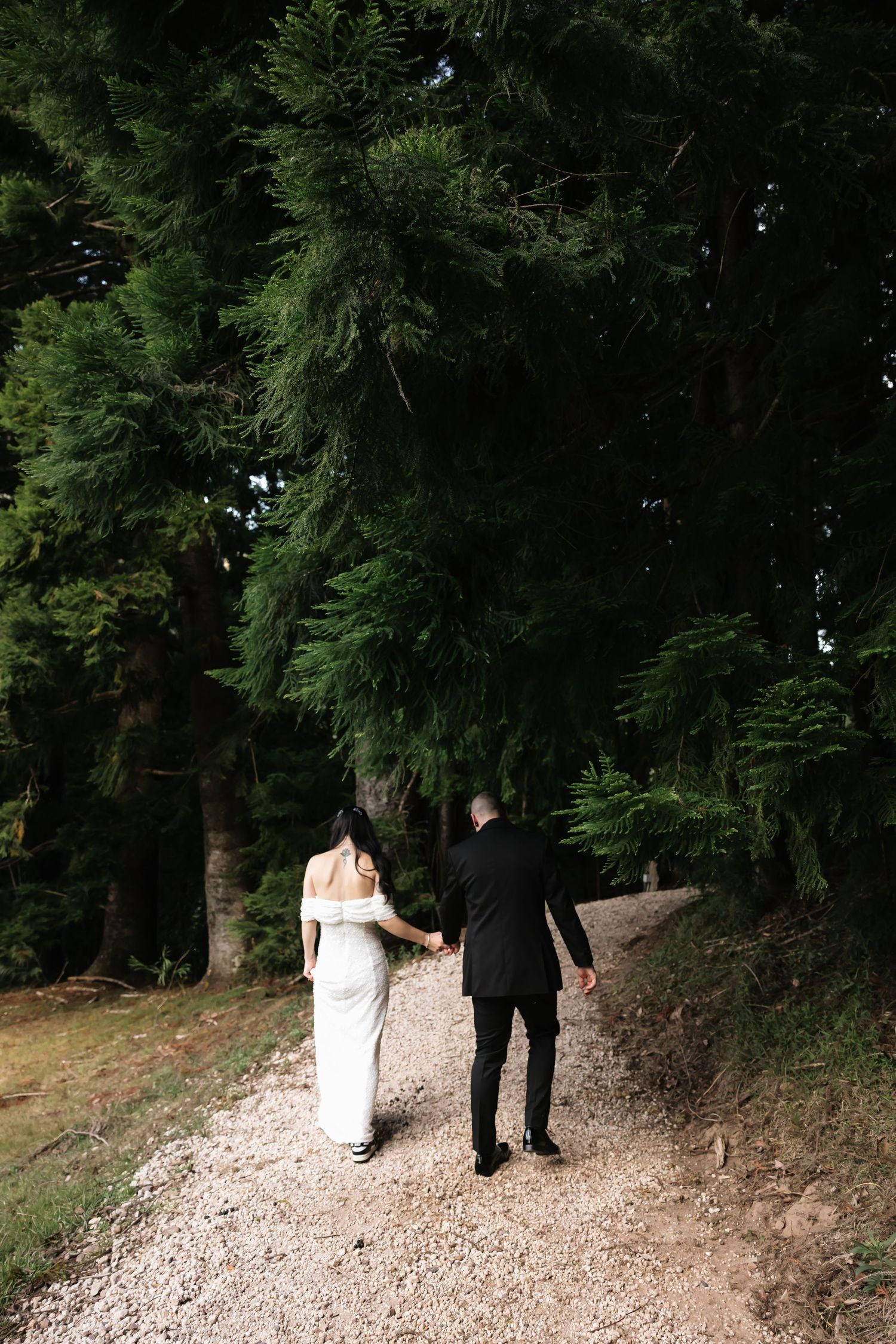 Bride in white gown and groom in black suit walking hand in hand along a forest gravel path.