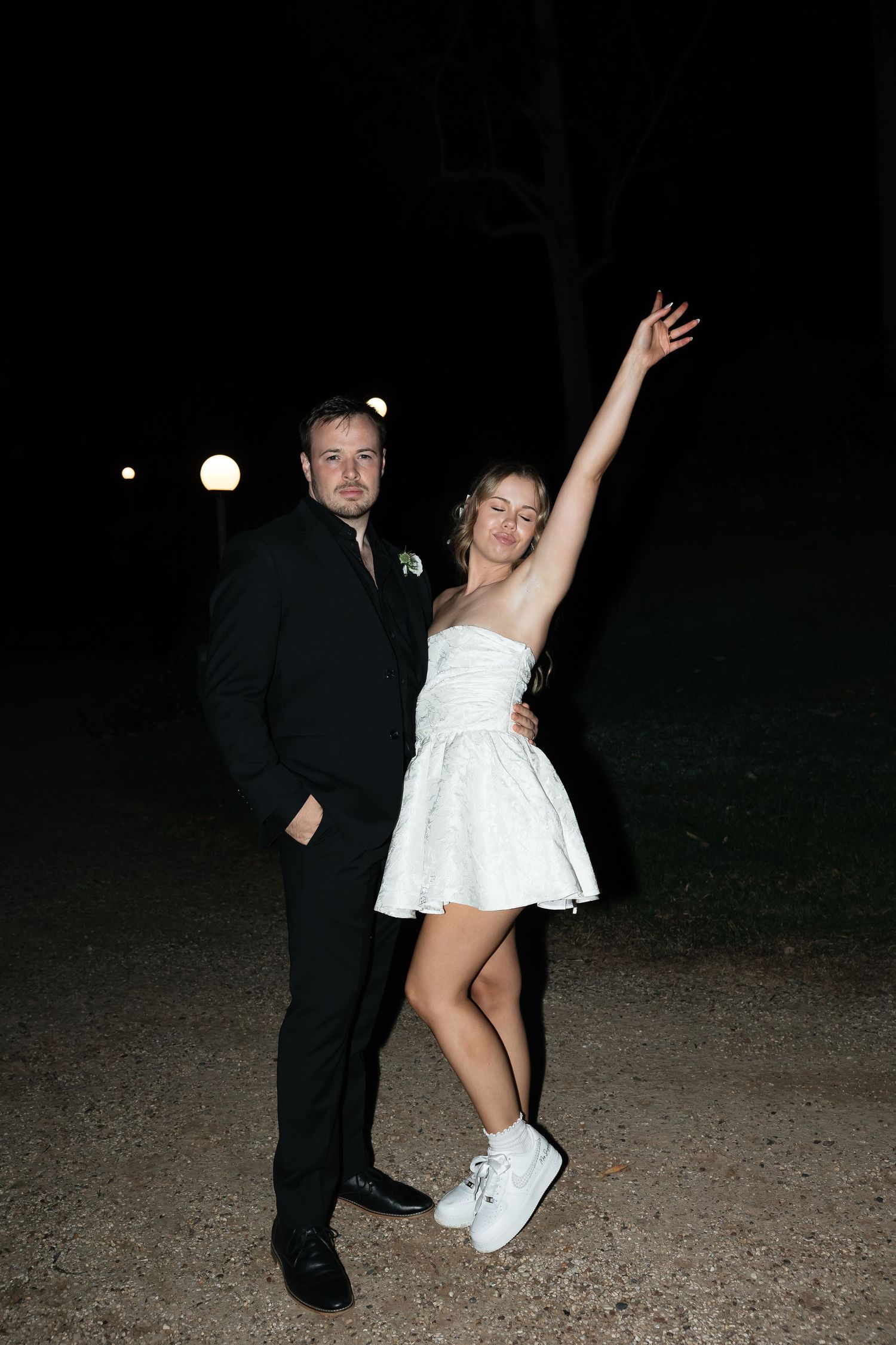 Newlywed couple celebrating at night, bride in short white dress and sneakers raising arm joyfully outdoors.