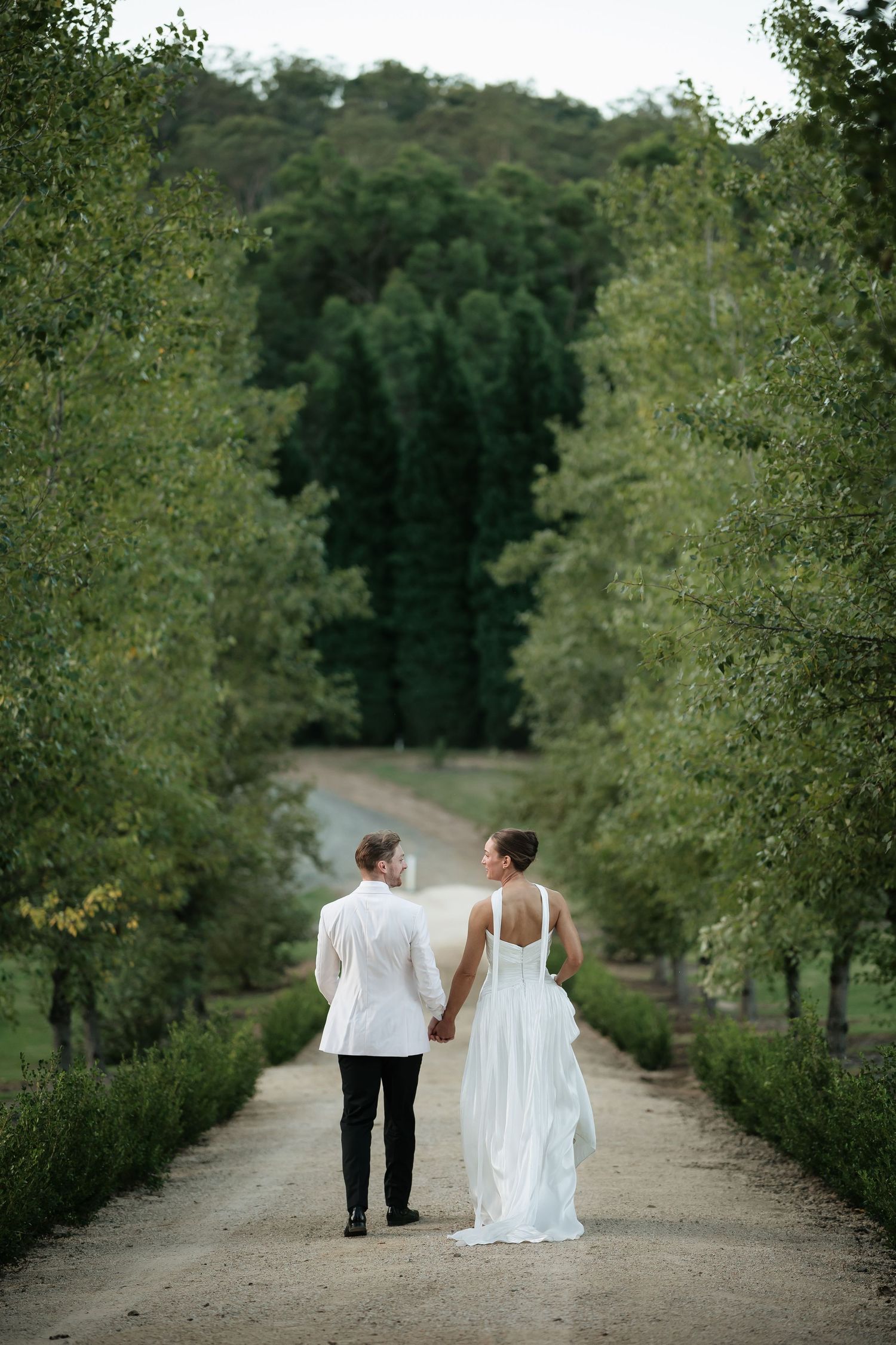 Bride and groom walking hand in hand down tree-lined gravel path surrounded by lush green gardens at The Woodhouse, Wollombi.