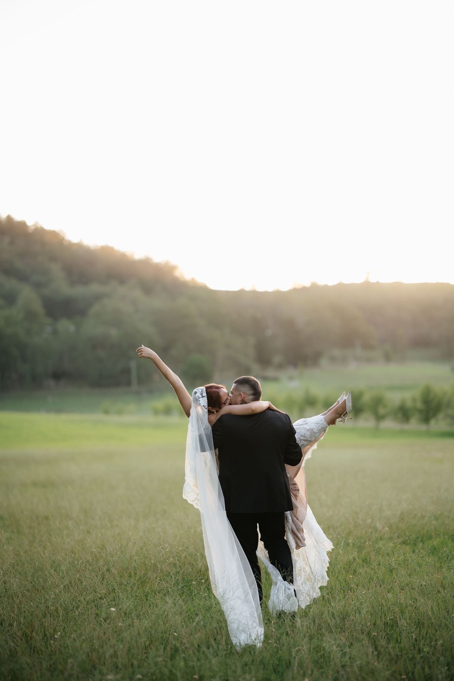 Groom lifts bride in a flowing lace wedding dress as she raises her arm joyfully in a sunlit field at Mystwood in the Hunter Valley.