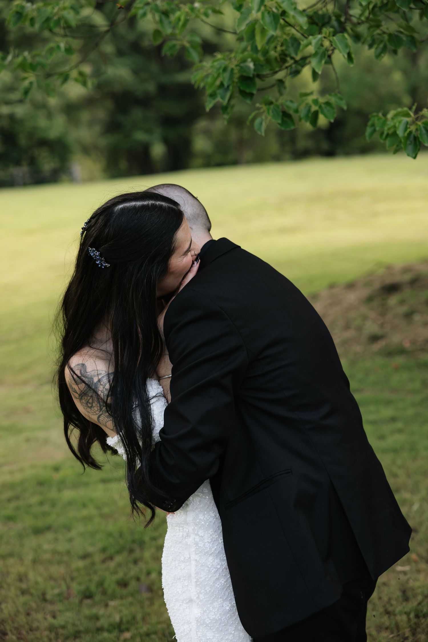 Bride with long dark hair and tattoos shares a kiss with groom in black suit outdoors.