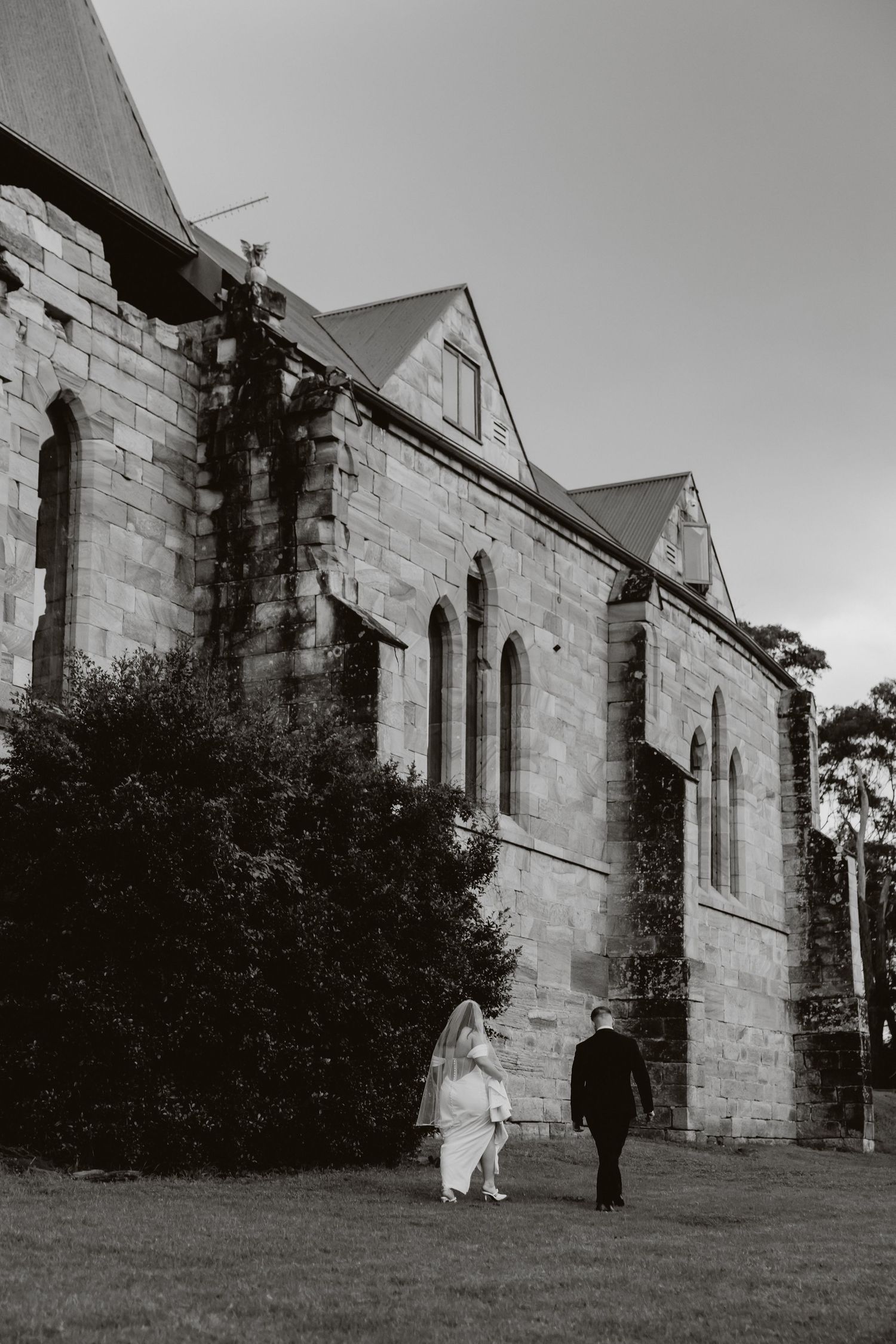 Black and white photo of bride and groom walking beside a historic stone church on their wedding day at St Joseph's Cathedral.