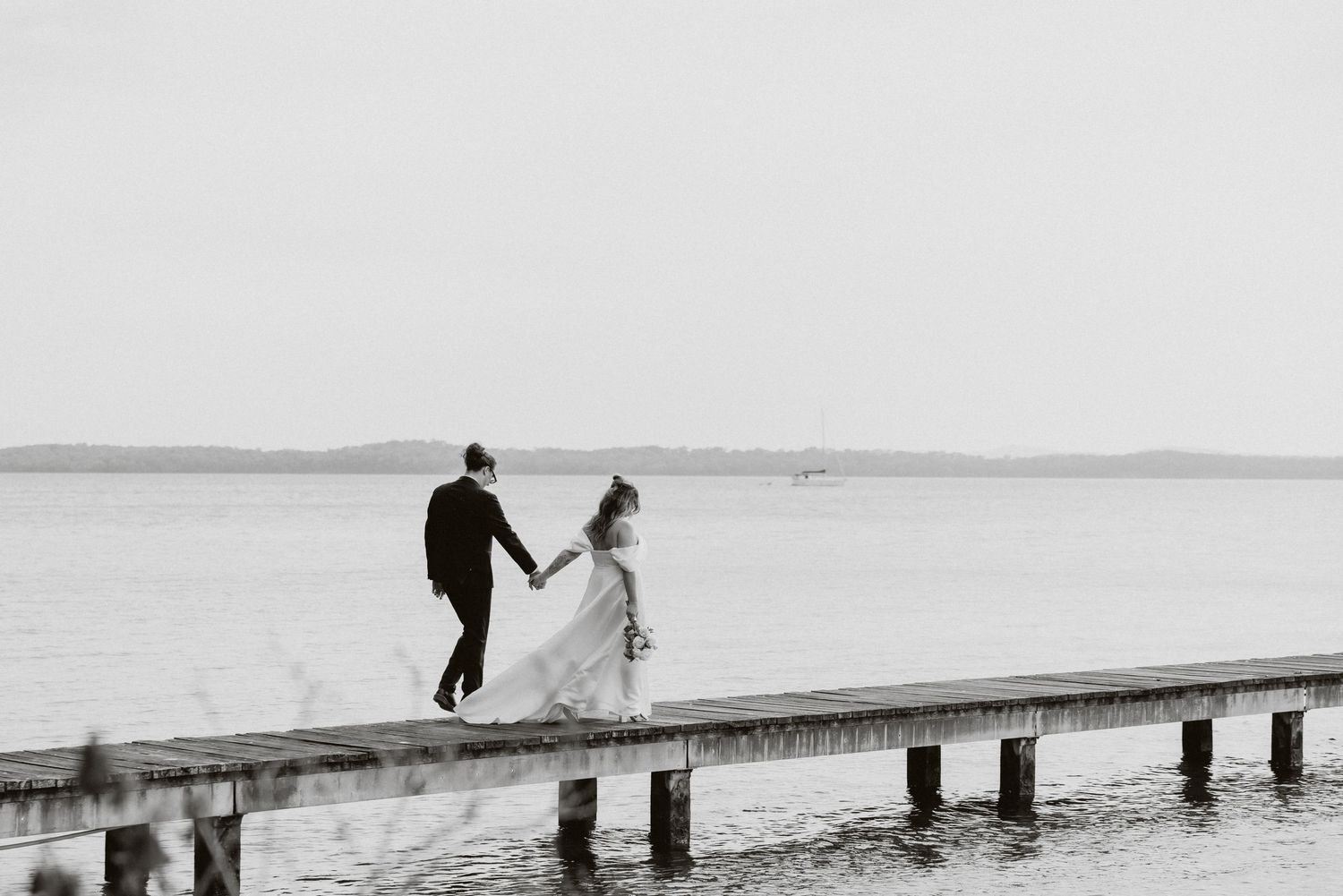 Black and white photo of bride and groom walking hand in hand on a waterfront pier.