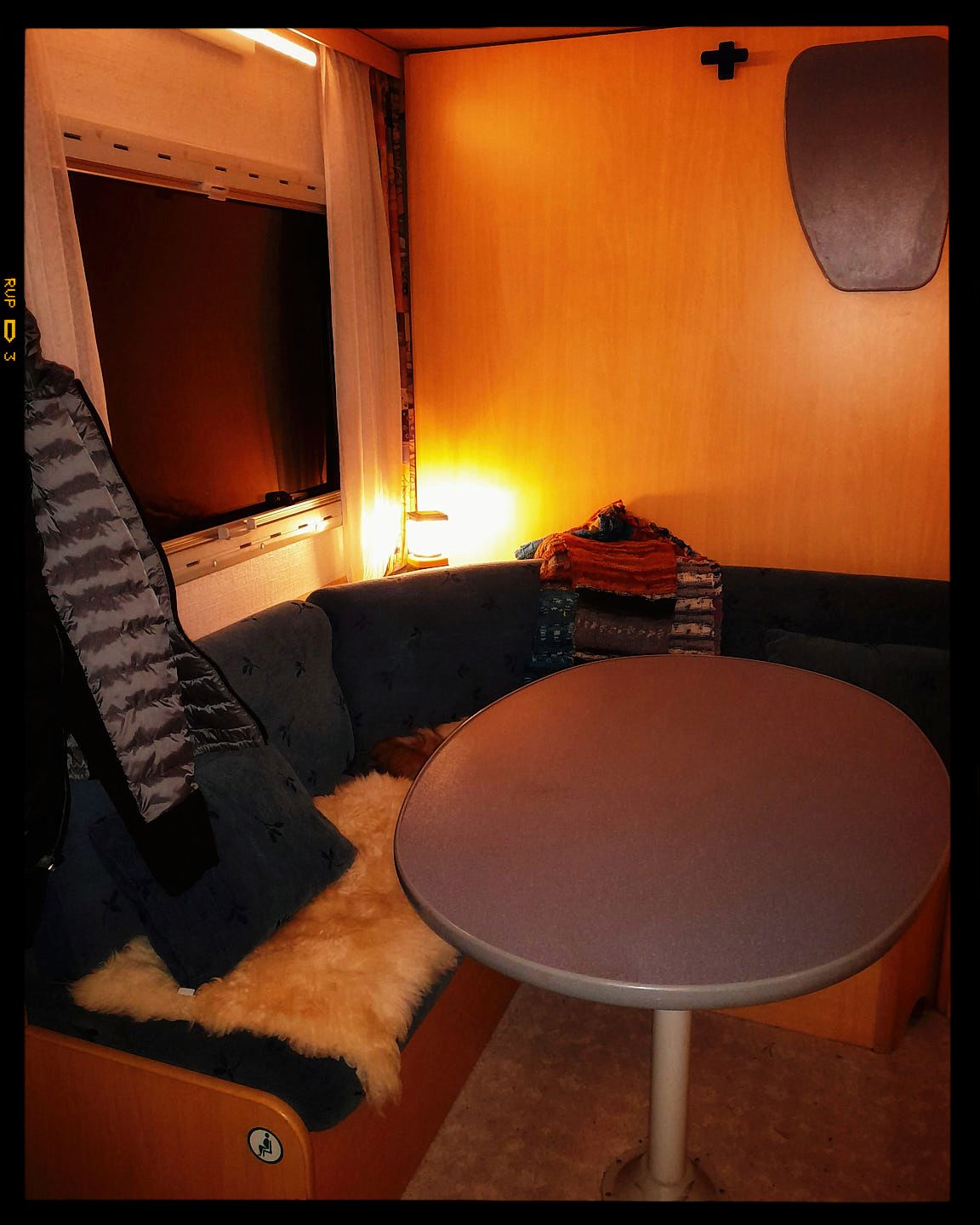 A cozy camper interior with warm lighting showing a round table, bench seating, and plush cushions against orange walls.