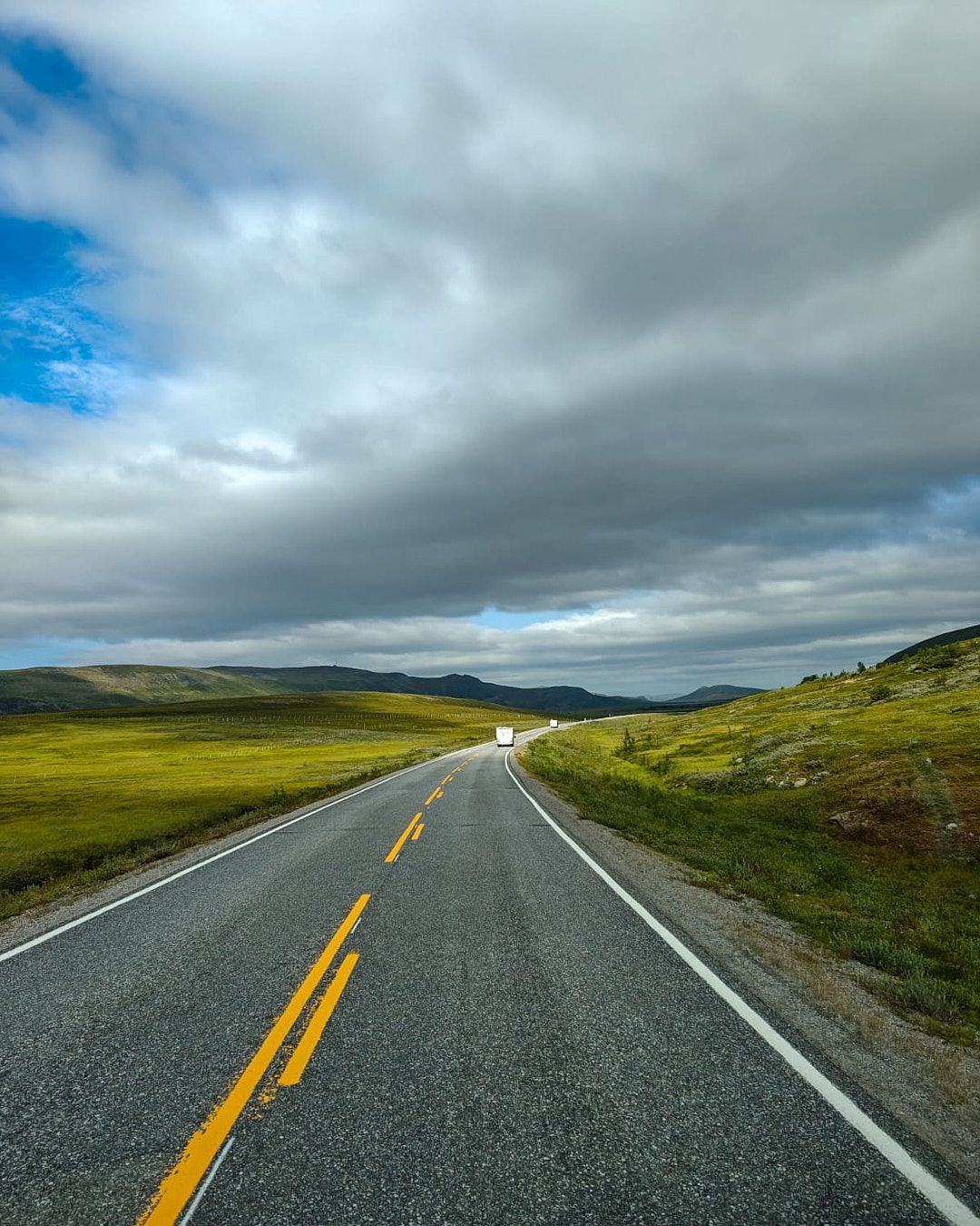 Empty rural highway with yellow lines stretches through rolling green hills under dramatic cloudy skies.