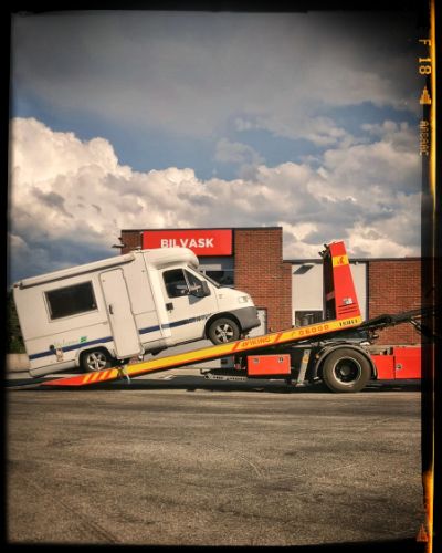 Four sequential photos showing a white RV being towed onto a flatbed truck in front of a brick building with a cloudy sky.