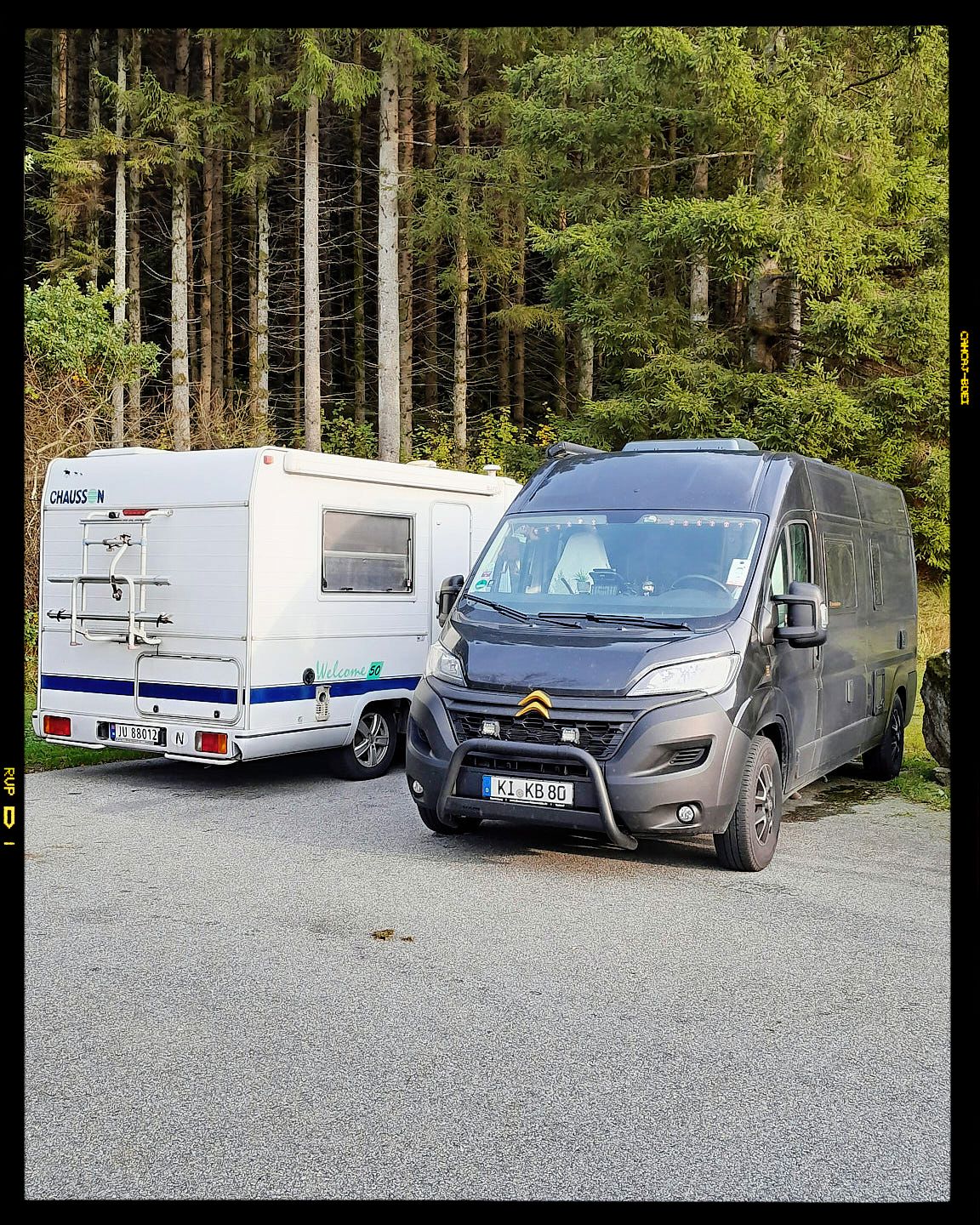 A blue van and white RV camper trailer parked in front of a dense forest of evergreen trees.