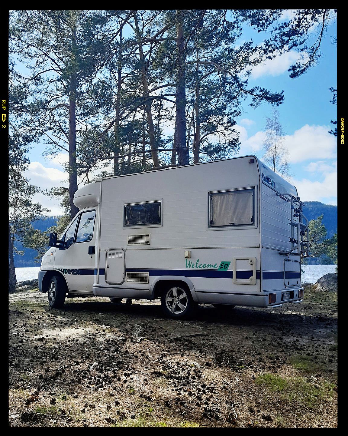 A white RV camper van parked among pine trees on a mountainside overlooking a scenic lake on a sunny day.