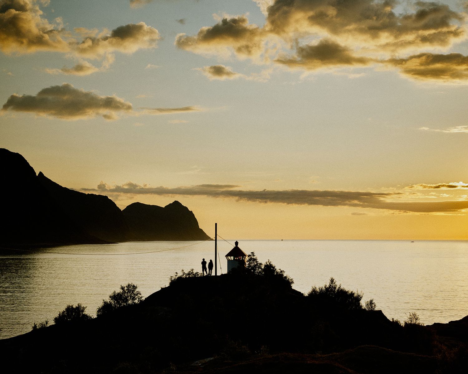 Silhouetted landscape at sunset showing coastal mountains and buildings against an orange sky.