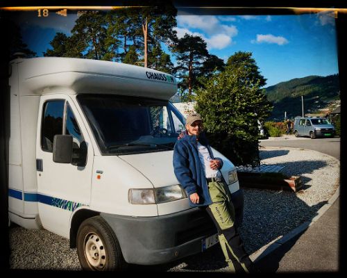 White camper van parked in gravel lot with mountains and pine trees in background on sunny day.