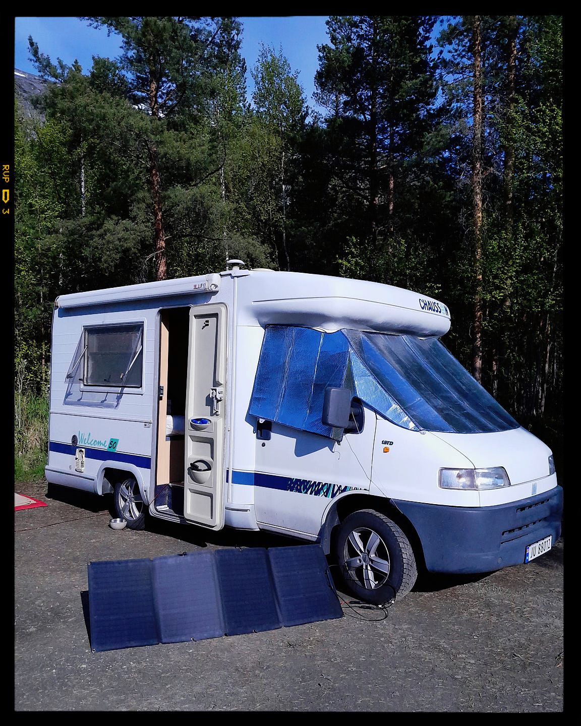 White motorhome camper van with solar panels displayed in front parked in a forested area on a sunny day.