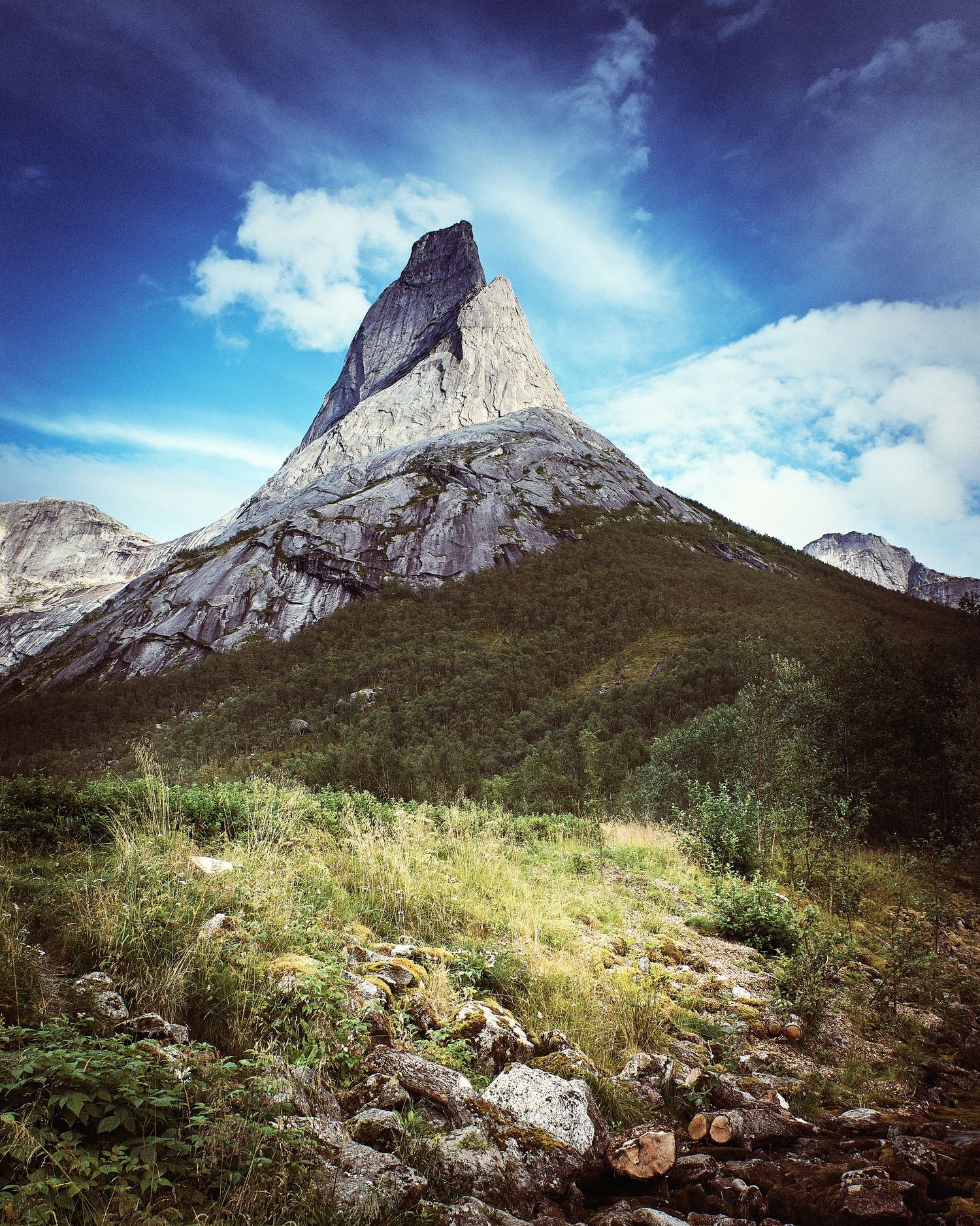Majestic mountain peak rises against blue sky with green slopes and rocky terrain in foreground.