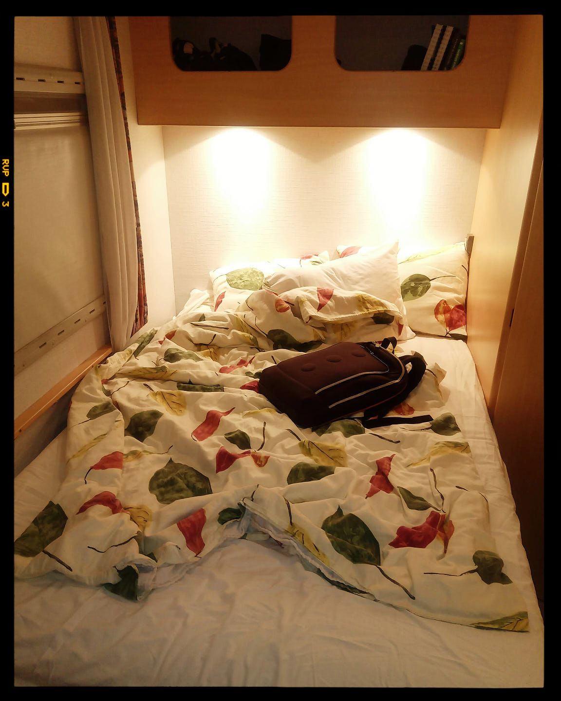 Unmade bed with floral patterned bedding and laptop under warm lighting in small sleeping compartment.