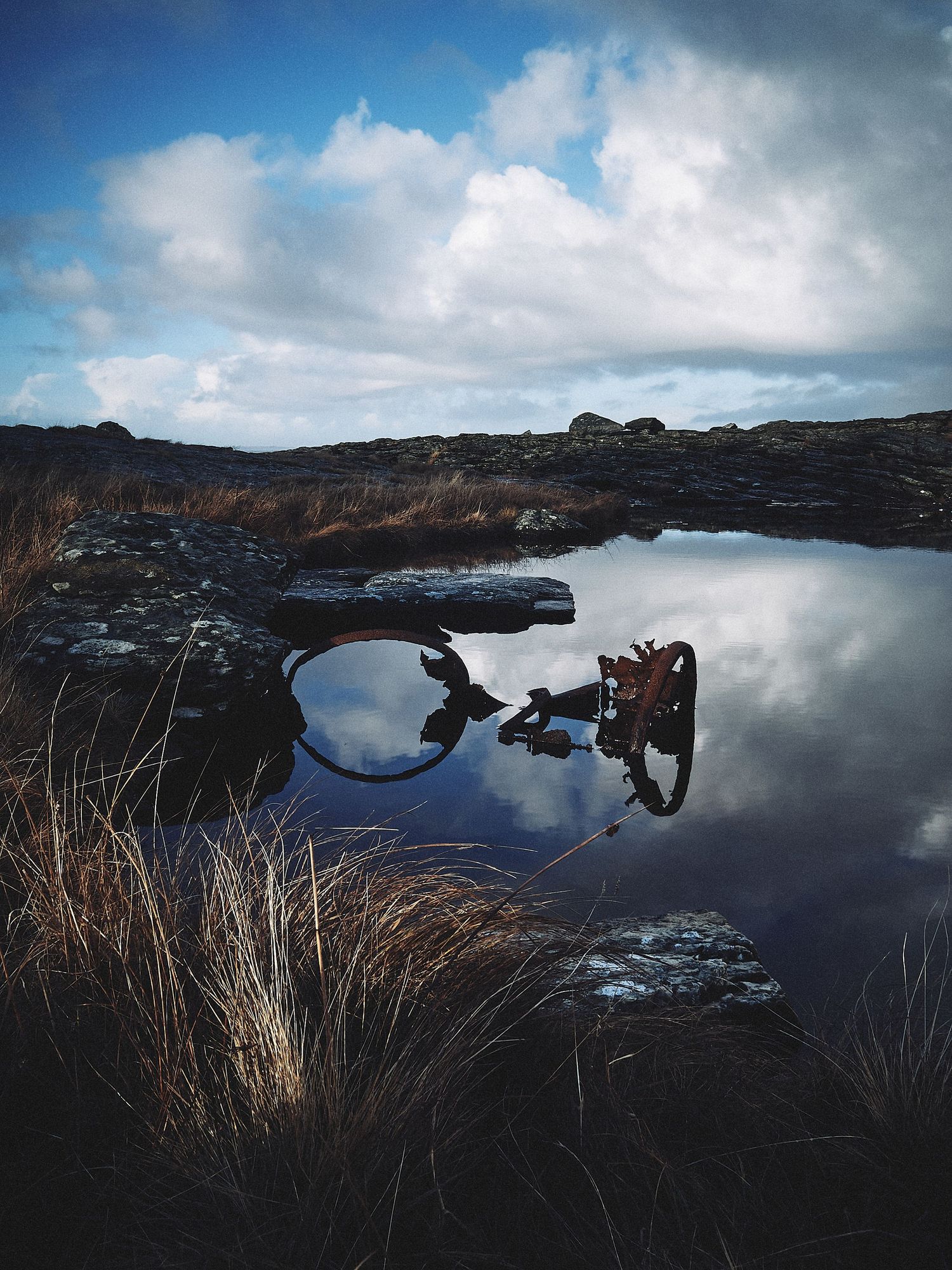 Reflective lake surface mirroring cloudy skies surrounded by dark rocky terrain and dried grass.