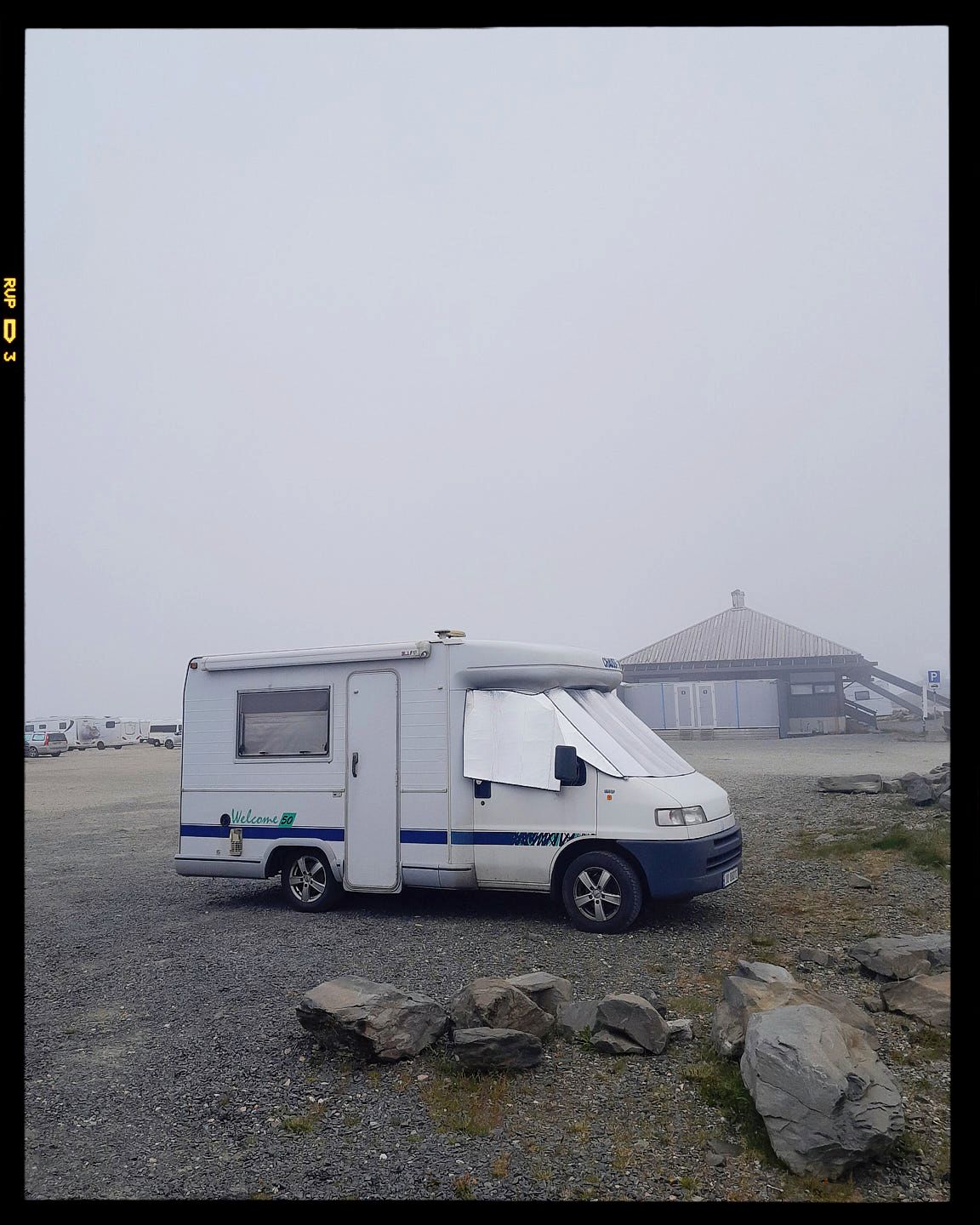A white camper van parked on rocky terrain in foggy conditions with a building silhouette in the background.