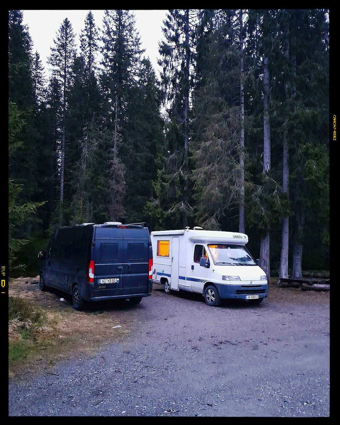 Several vehicles and camper trailers parked in a gravel lot surrounded by tall evergreen trees in a forest setting.