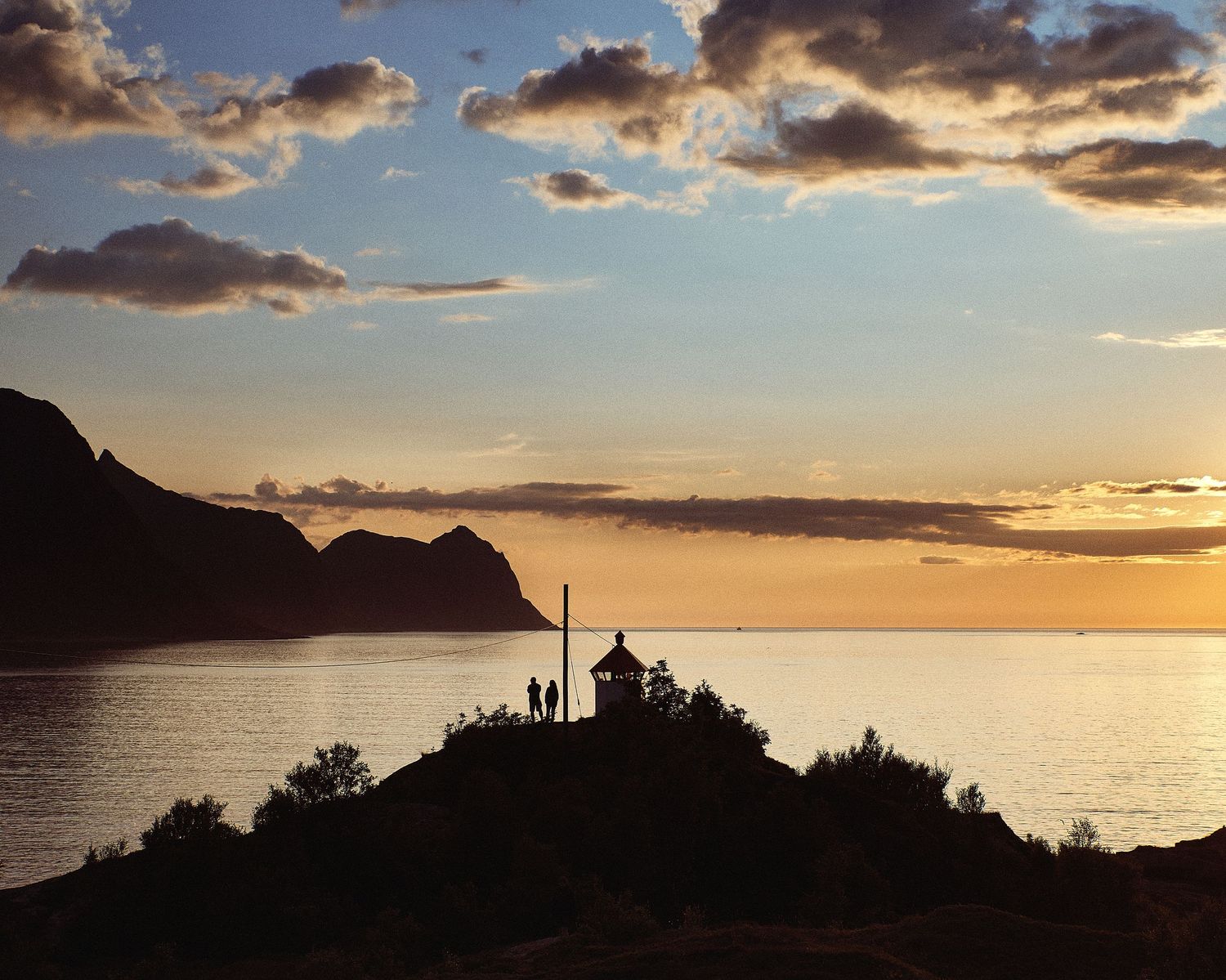 Coastal landscape silhouette at sunset with dramatic orange clouds and mountain outlines.