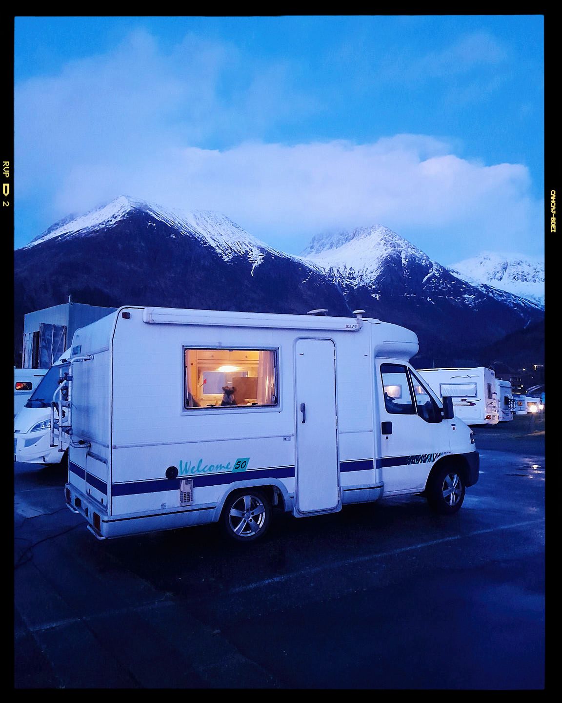 A white RV camper parked at dusk with snow-capped mountains and blue sky in the background, warm light glowing from window.
