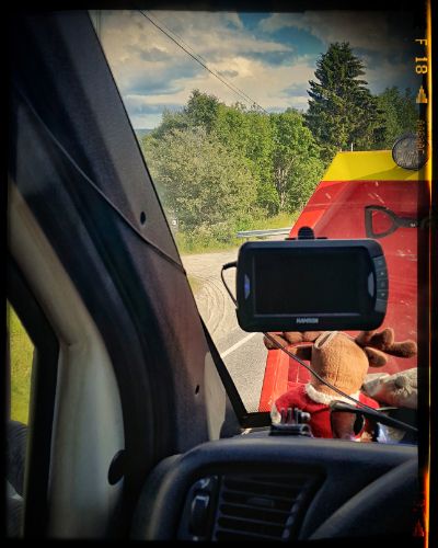 View through car windshield of a red pickup truck or tractor on a rural road with trees and cloudy sky in background.