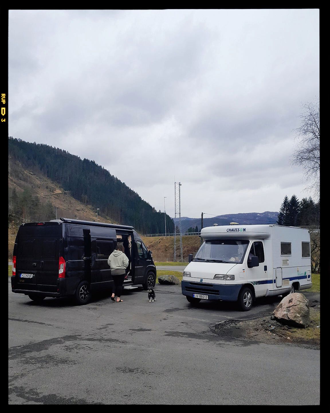 Three white RV campers and dark vehicles parked in a gravel lot with mountains and cloudy skies in the background.