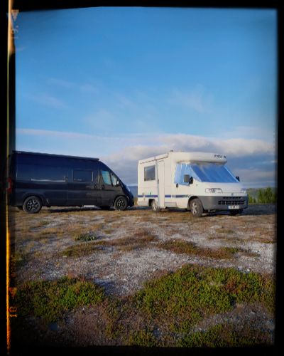 A group of recreational vehicles and cars parked on a rugged coastal terrain under a bright blue sky.
