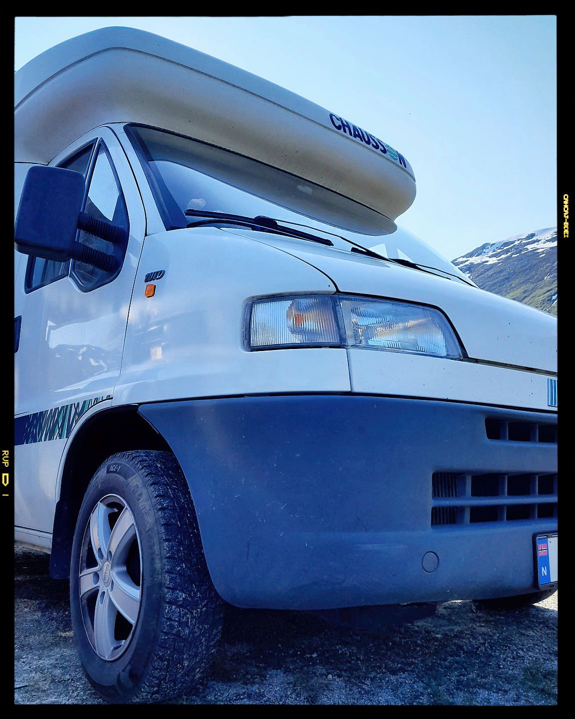 A white and blue motorhome with rooftop cargo box parked in a snowy mountain landscape.
