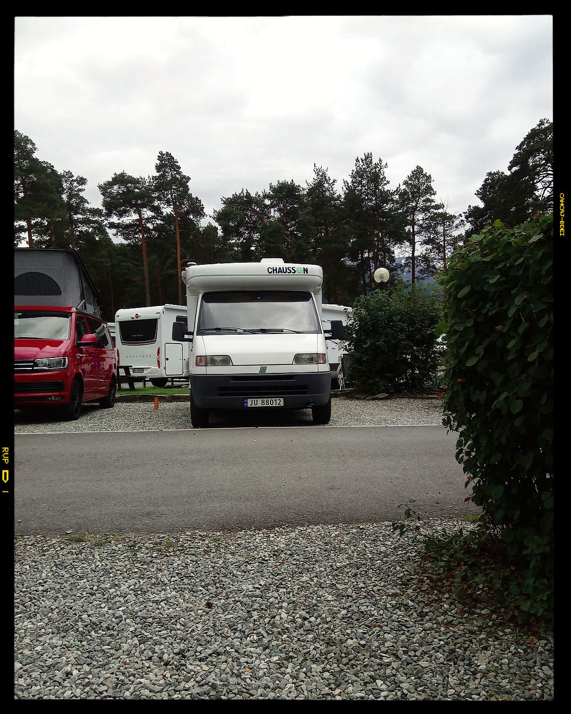 A white motorhome and red car parked in a gravel lot surrounded by tall pine trees on an overcast day.