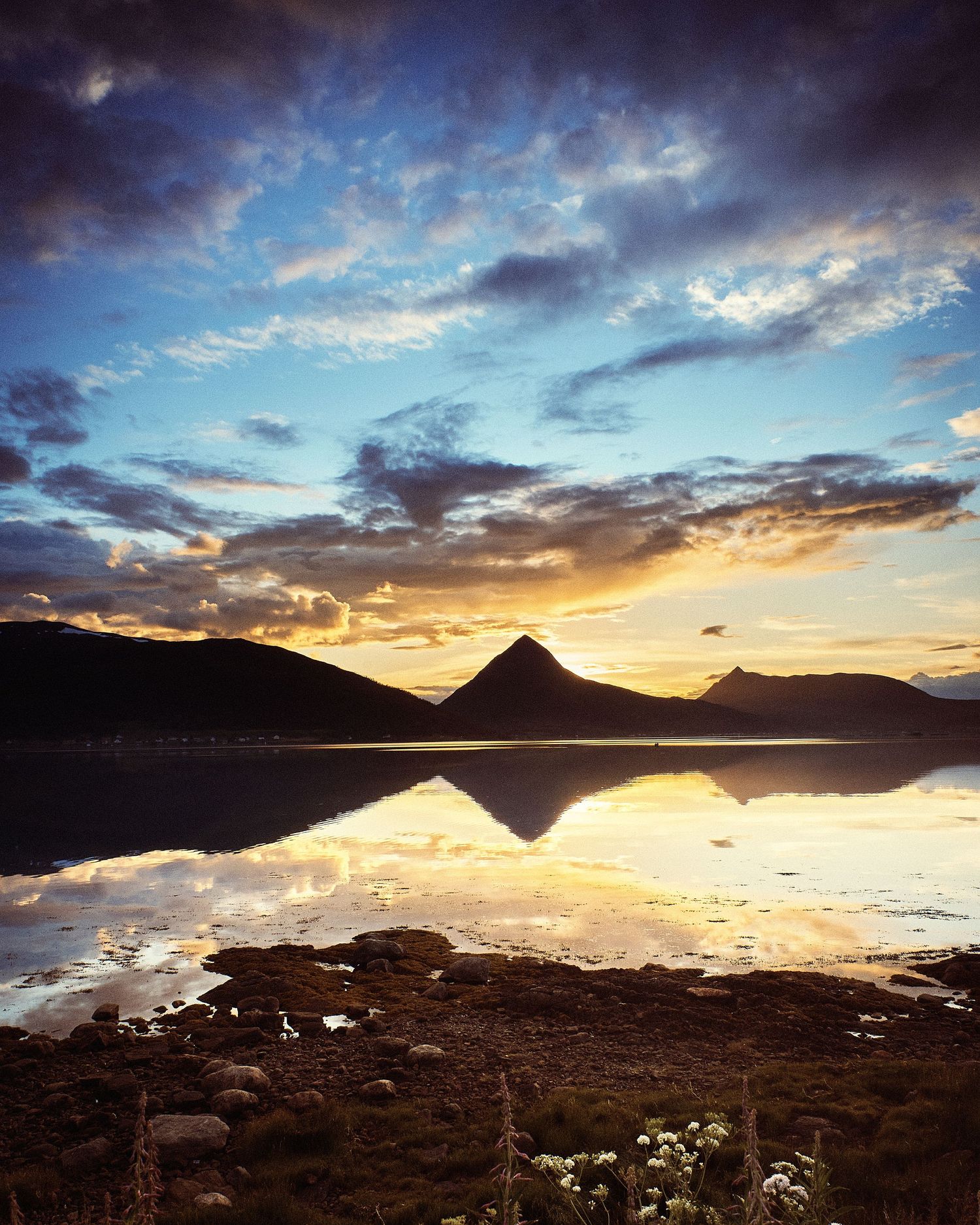 Dramatic sunset skyline reflected in still waters with mountain silhouettes and rocky shoreline.