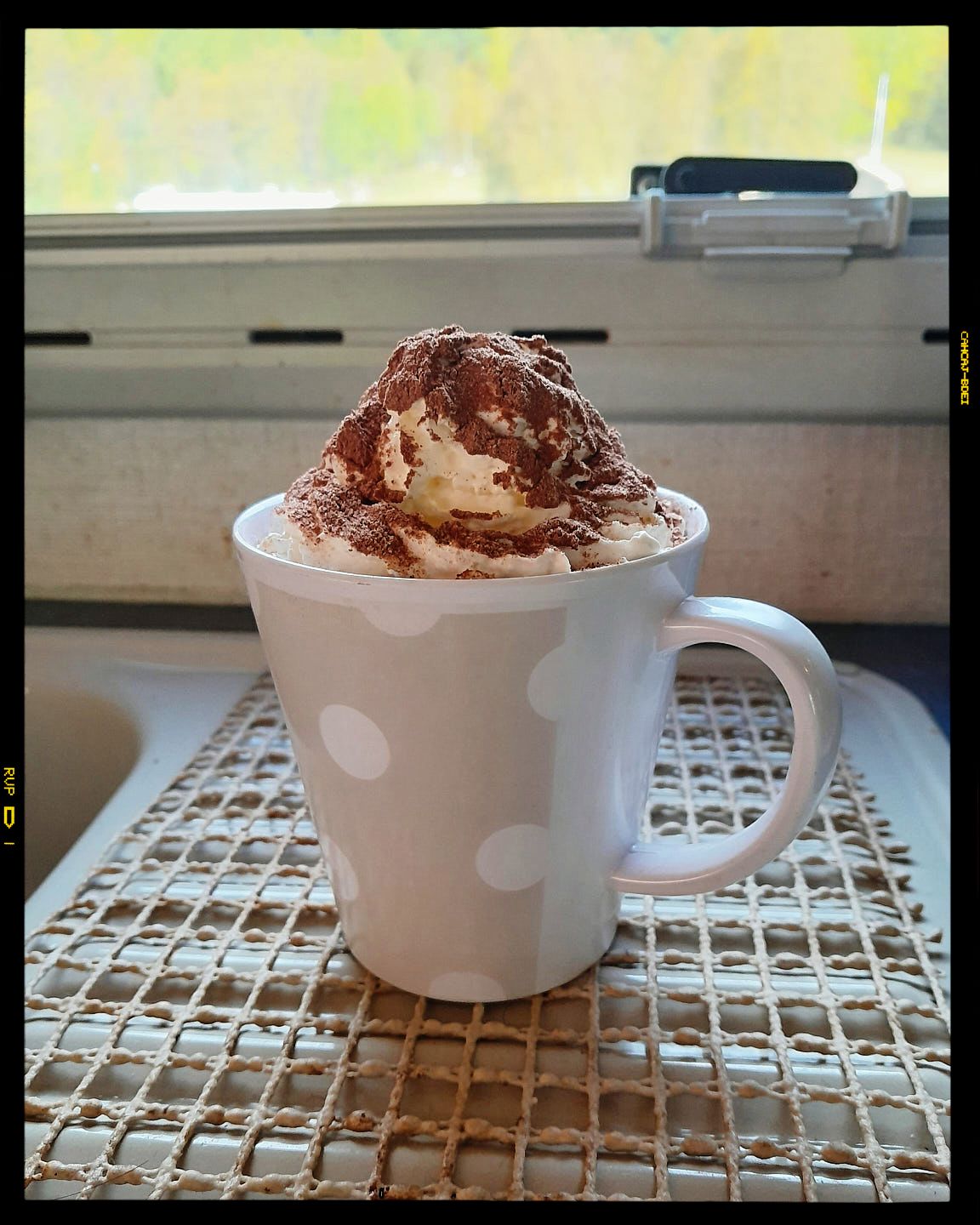 A white polka dot mug filled with whipped cream and chocolate shavings sits on a woven placemat.