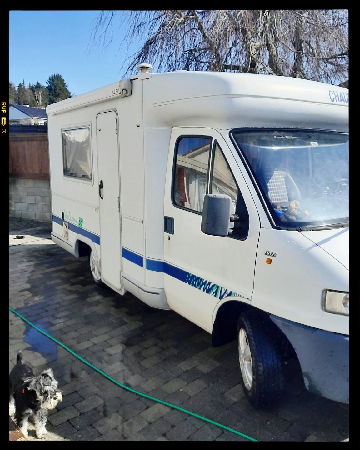 White motorhome with blue accent stripes parked in a driveway with a green garden hose visible.