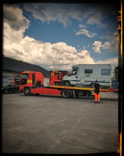 A red flatbed tow truck with a long trailer parked in an open parking lot under cloudy skies at dusk.