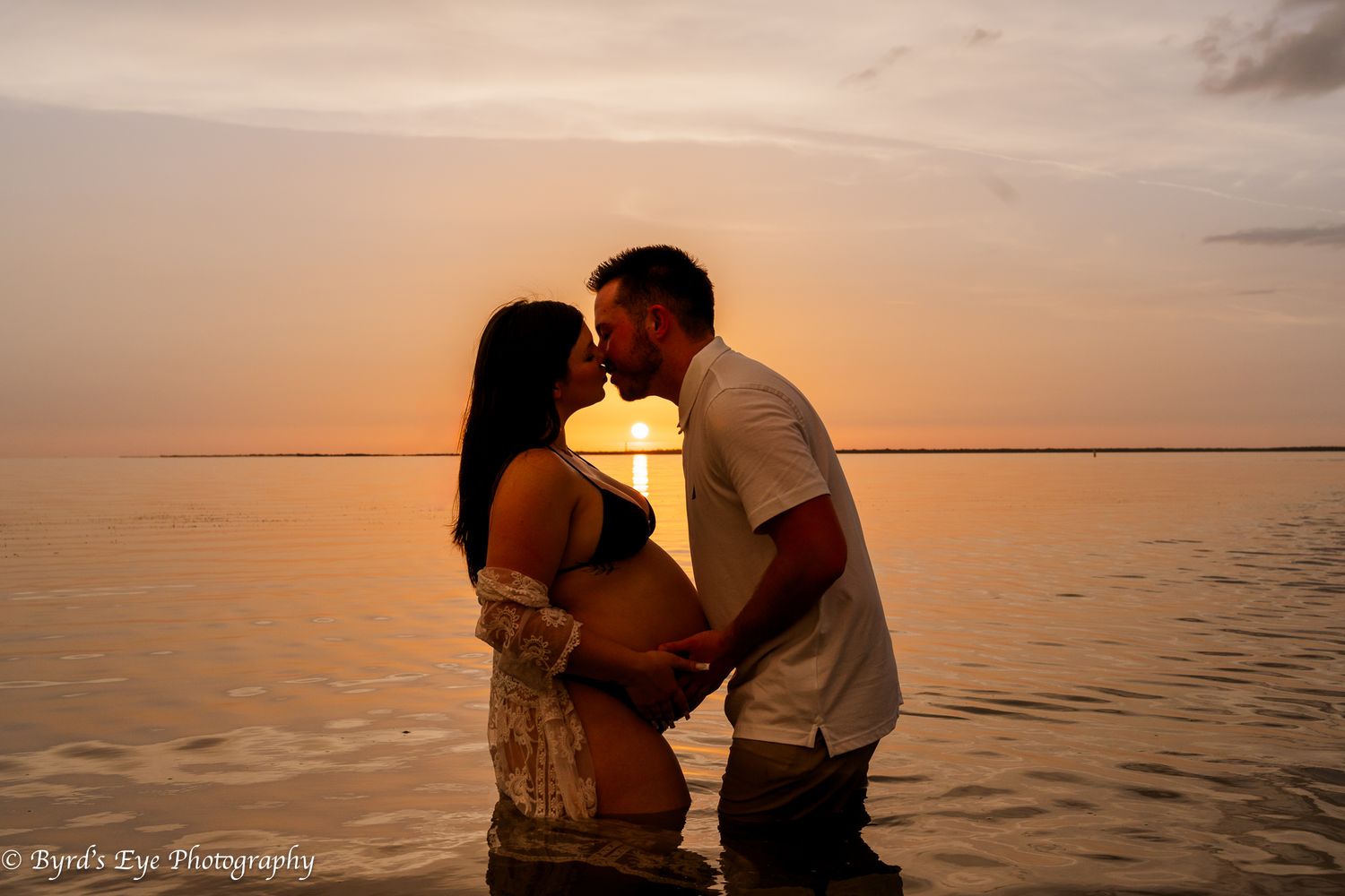 A couple shares a romantic moment during sunset while standing in calm ocean waters.