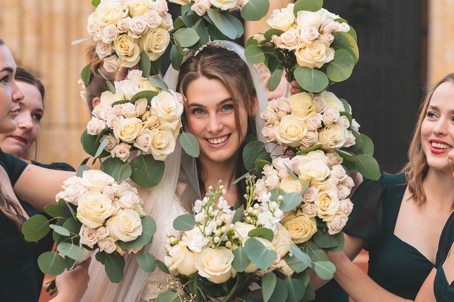 Wedding party holding cream-colored rose bouquets with eucalyptus leaves against a dark background.
