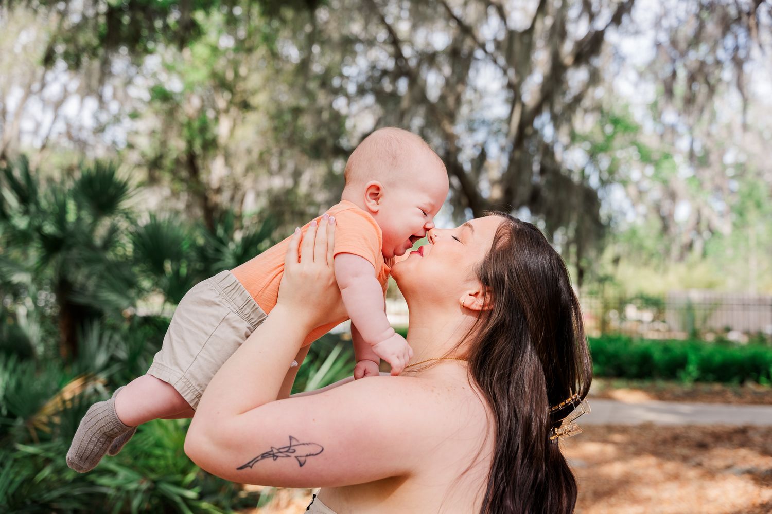 A parent lifts their baby up in a joyful moment together under trees in an outdoor garden setting.