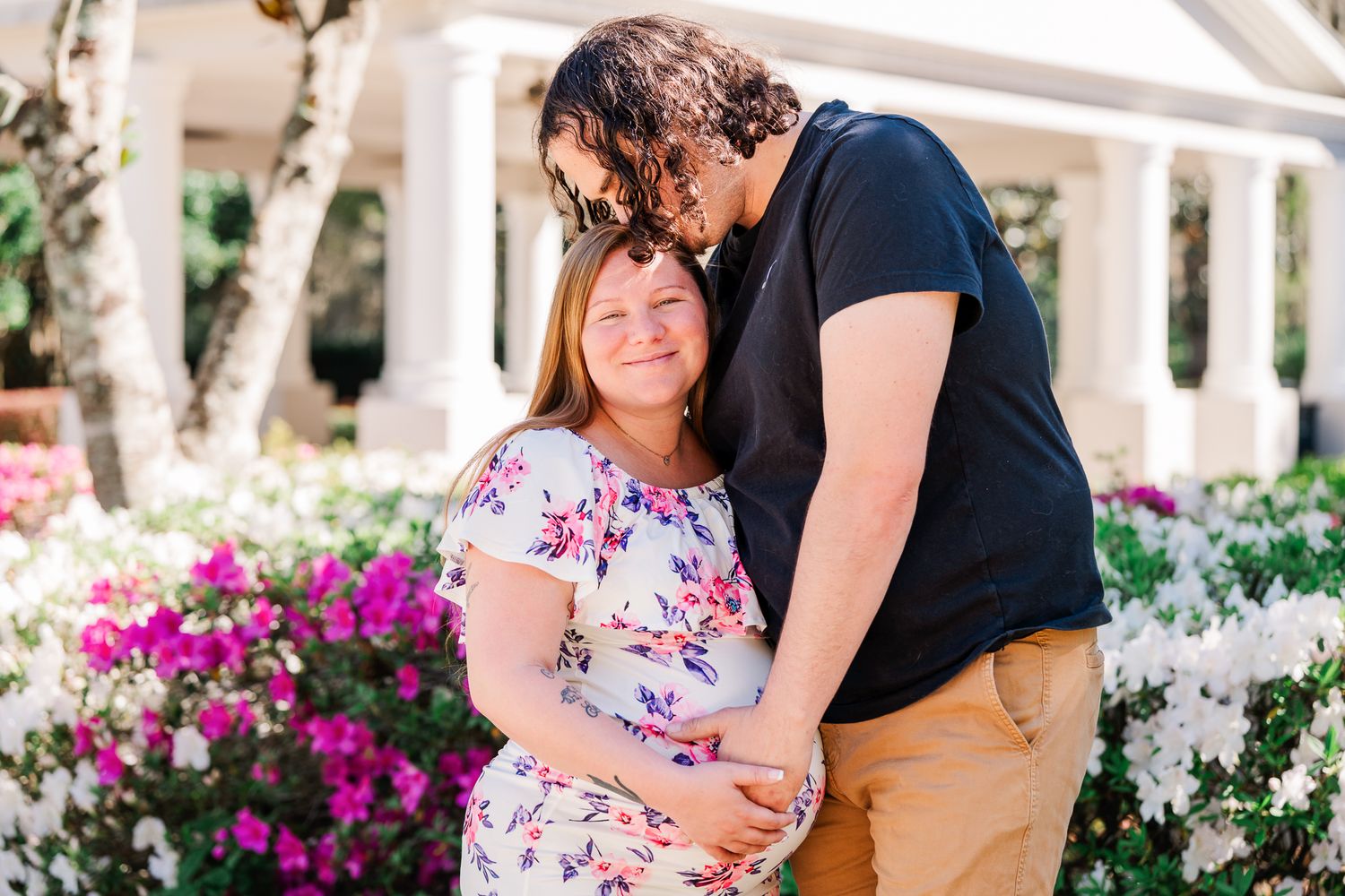 A loving couple shares a tender moment in a garden with blooming pink bougainvillea flowers and white columns.