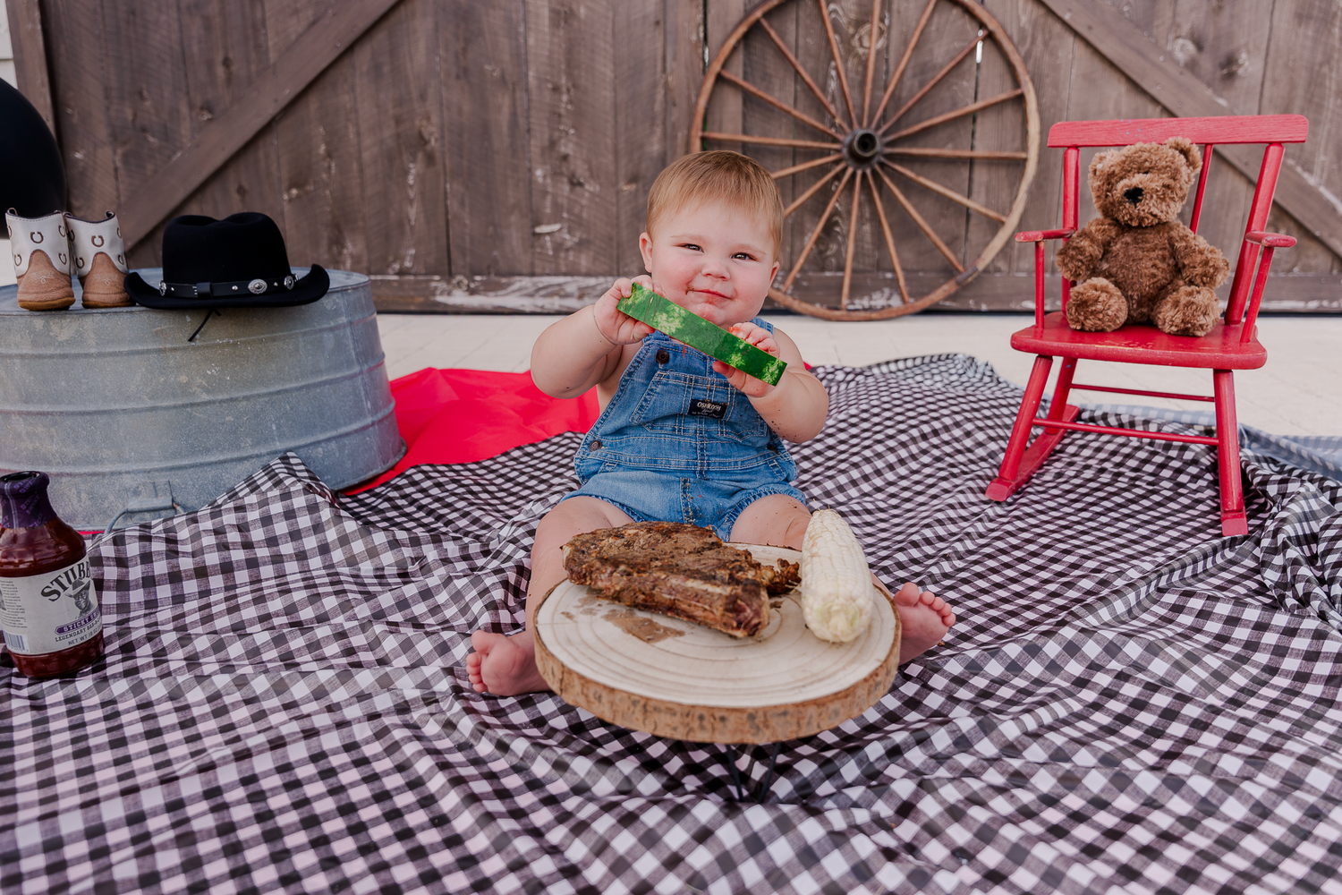 A baby eats cake while sitting on a checkered blanket with a wagon wheel and red chair in the background.