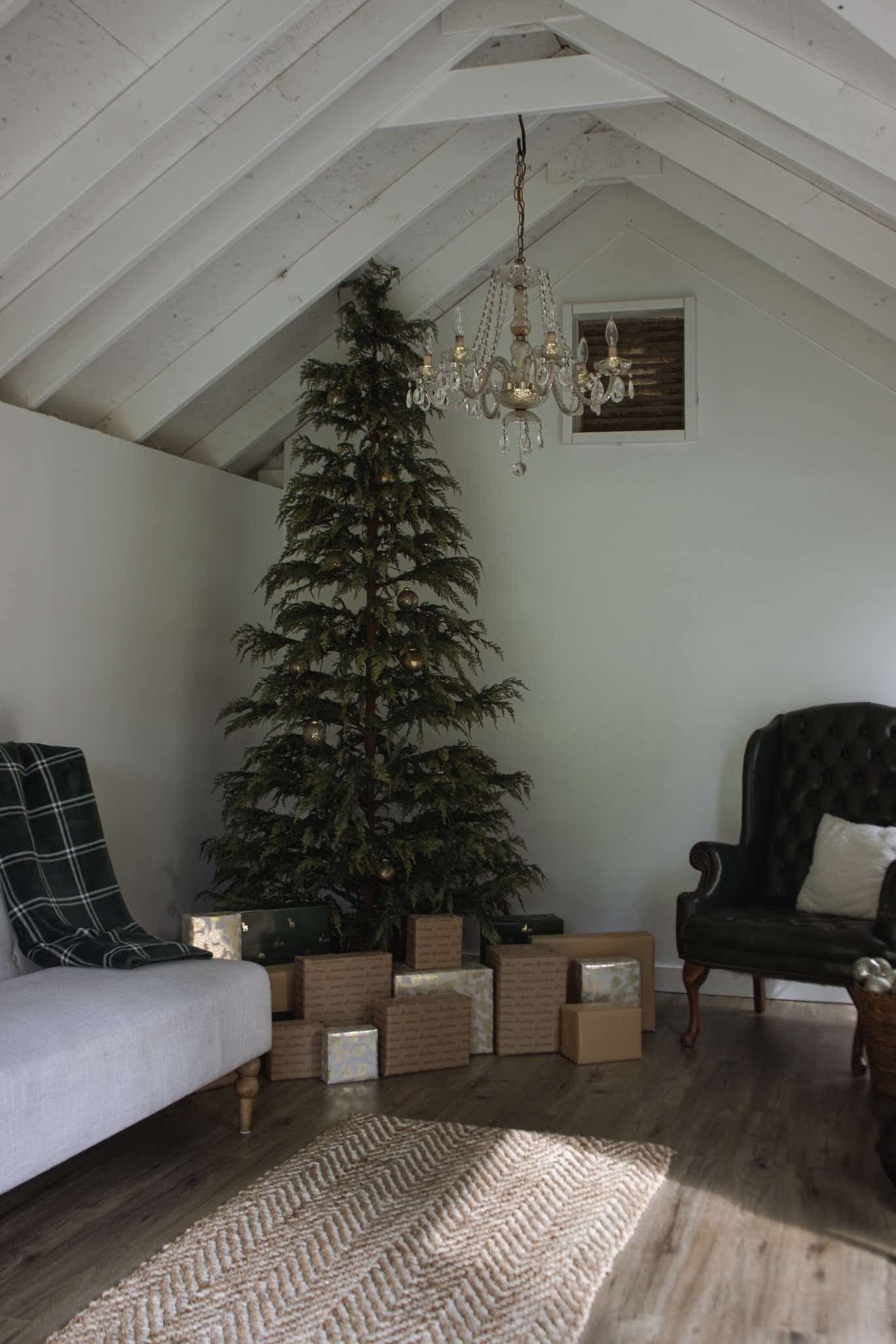 Cozy living room with tall decorated Christmas tree, wrapped gifts, crystal chandelier, and dark green armchair under vaulted ceiling.