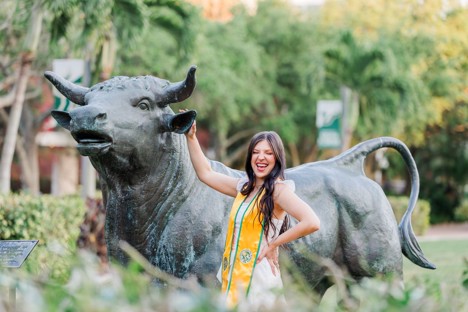 Graduate in yellow stole poses with bronze bull statue in garden setting.